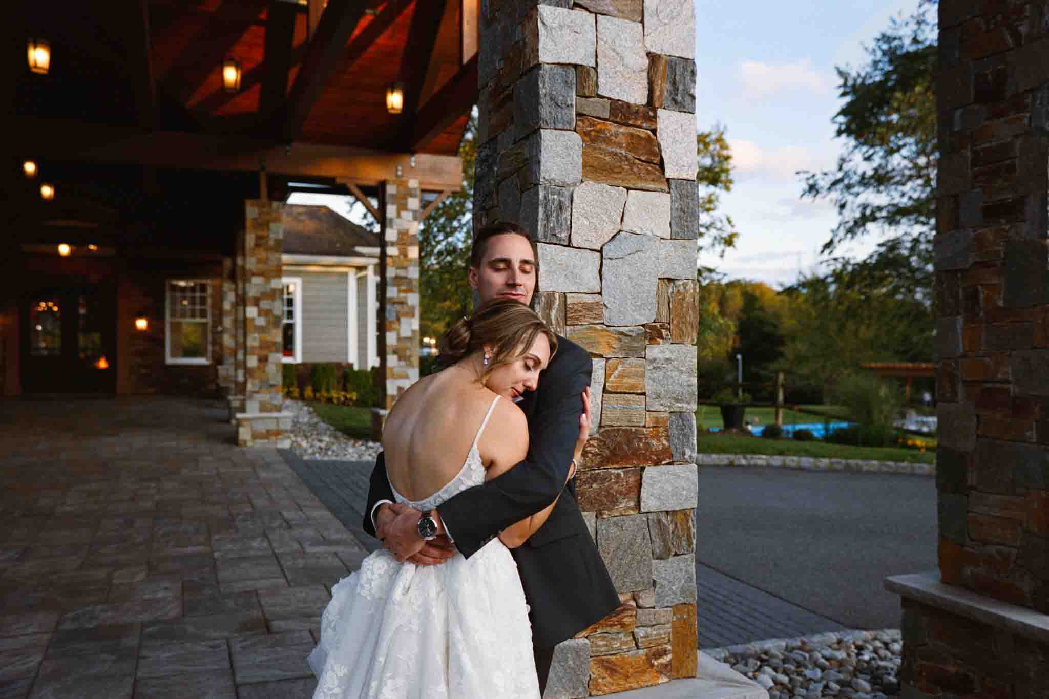 A bride in a white dress leans back, smiling, as she hugs her groom in a dark suit against a stone pillar outdoors at sunset—captured by a documentary wedding photographer specializing in candid film wedding photos in New Jersey.