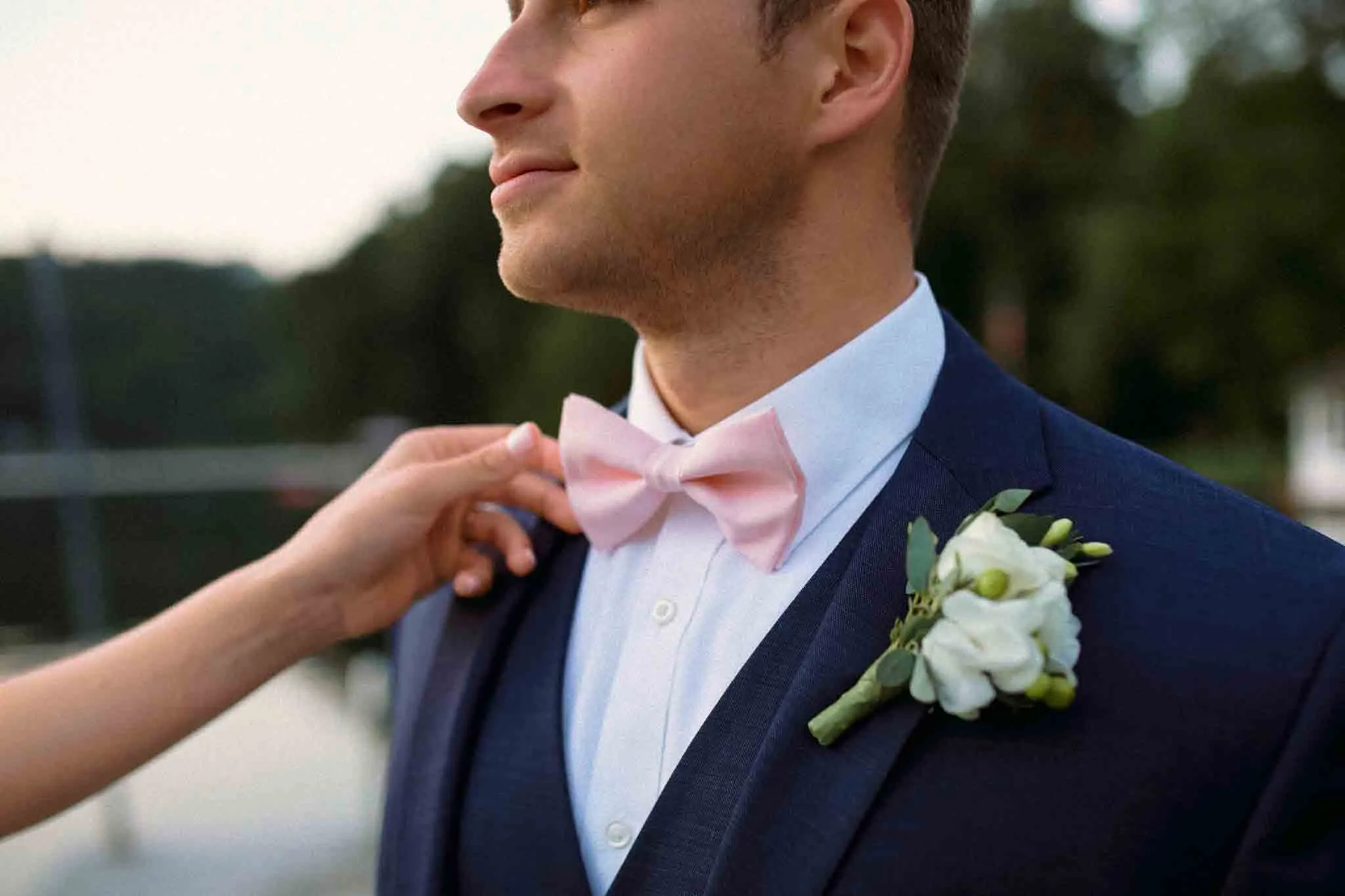 A close-up of a groom in a navy suit with a light pink bow tie and white boutonniere, as a hand adjusts his bow tie. Captured by a New Jersey film wedding photographer, the outdoor background is filled with trees and blurred scenery.