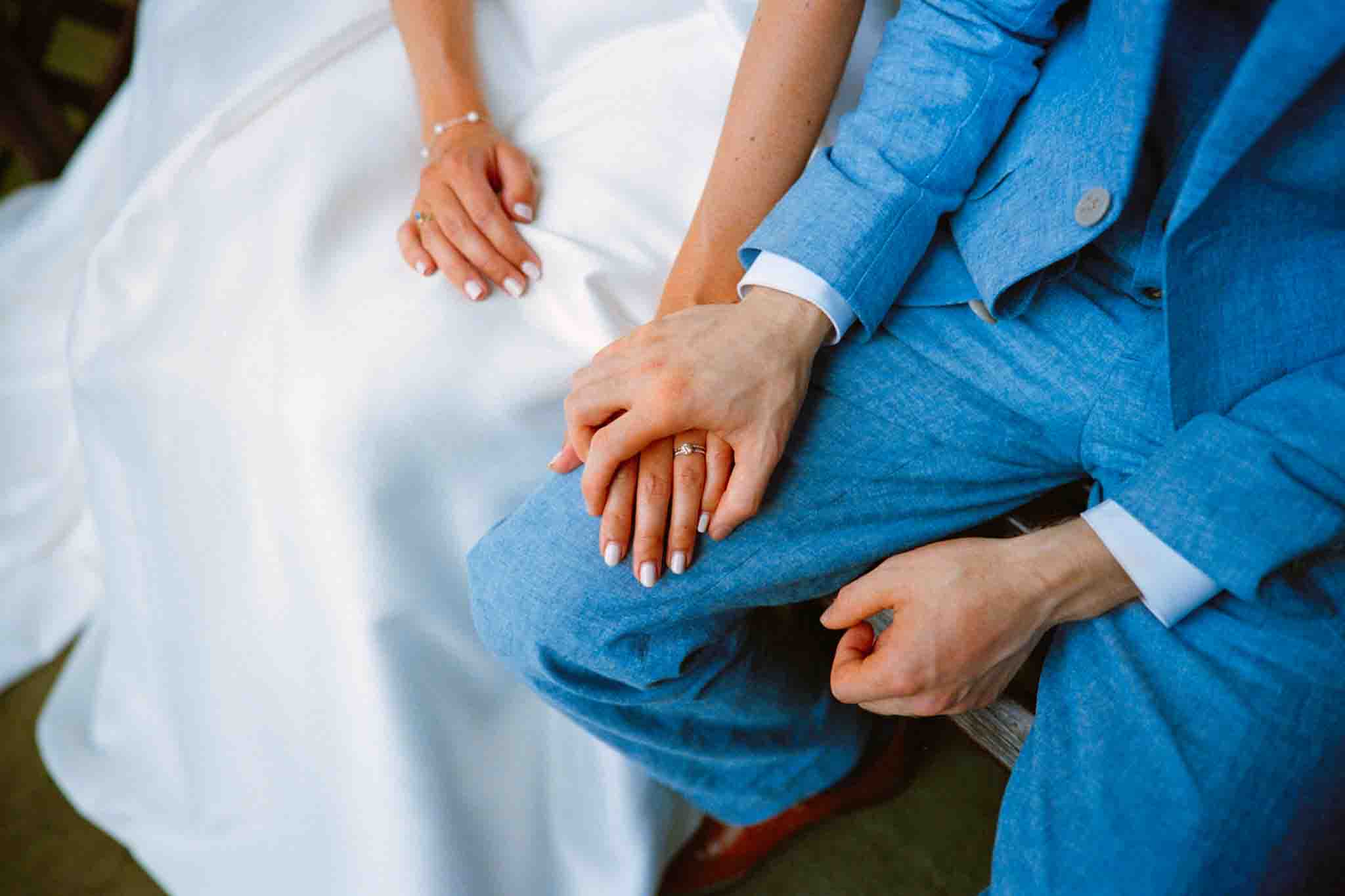 A close-up of a couple sitting side by side, wearing a white dress and a light blue suit. Their hands, adorned with wedding rings, gently rest together—an authentic moment captured in candid film wedding photos New Jersey couples treasure.