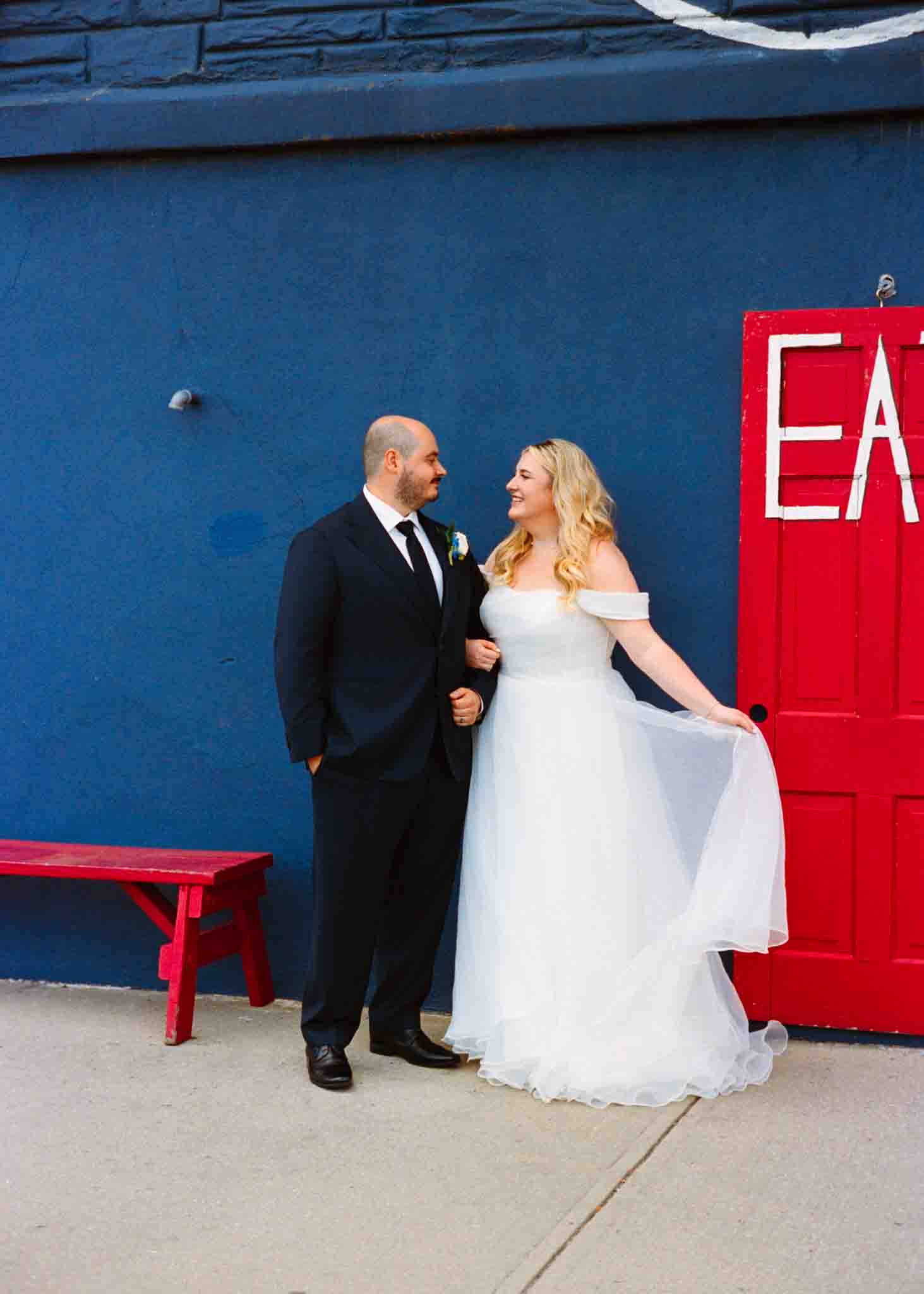 A bride in a white dress and a groom in a dark suit stand smiling at each other in front of a blue wall with a red door that says EAT and a red bench nearby, captured by an analog wedding photography NJ specialist.