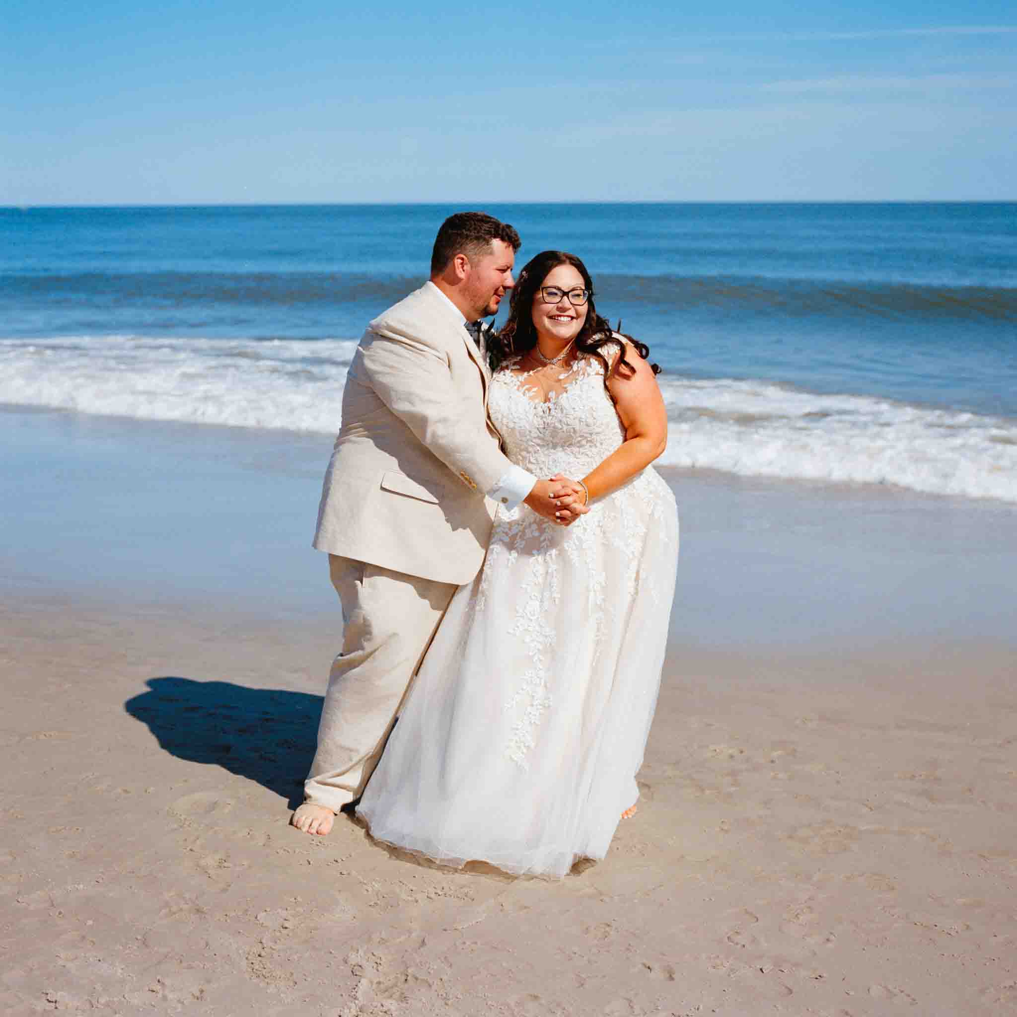 A smiling couple in wedding attire stands barefoot on a sandy beach, holding hands with ocean waves and a clear blue sky in the background—perfect for candid film wedding photos New Jersey couples will cherish.