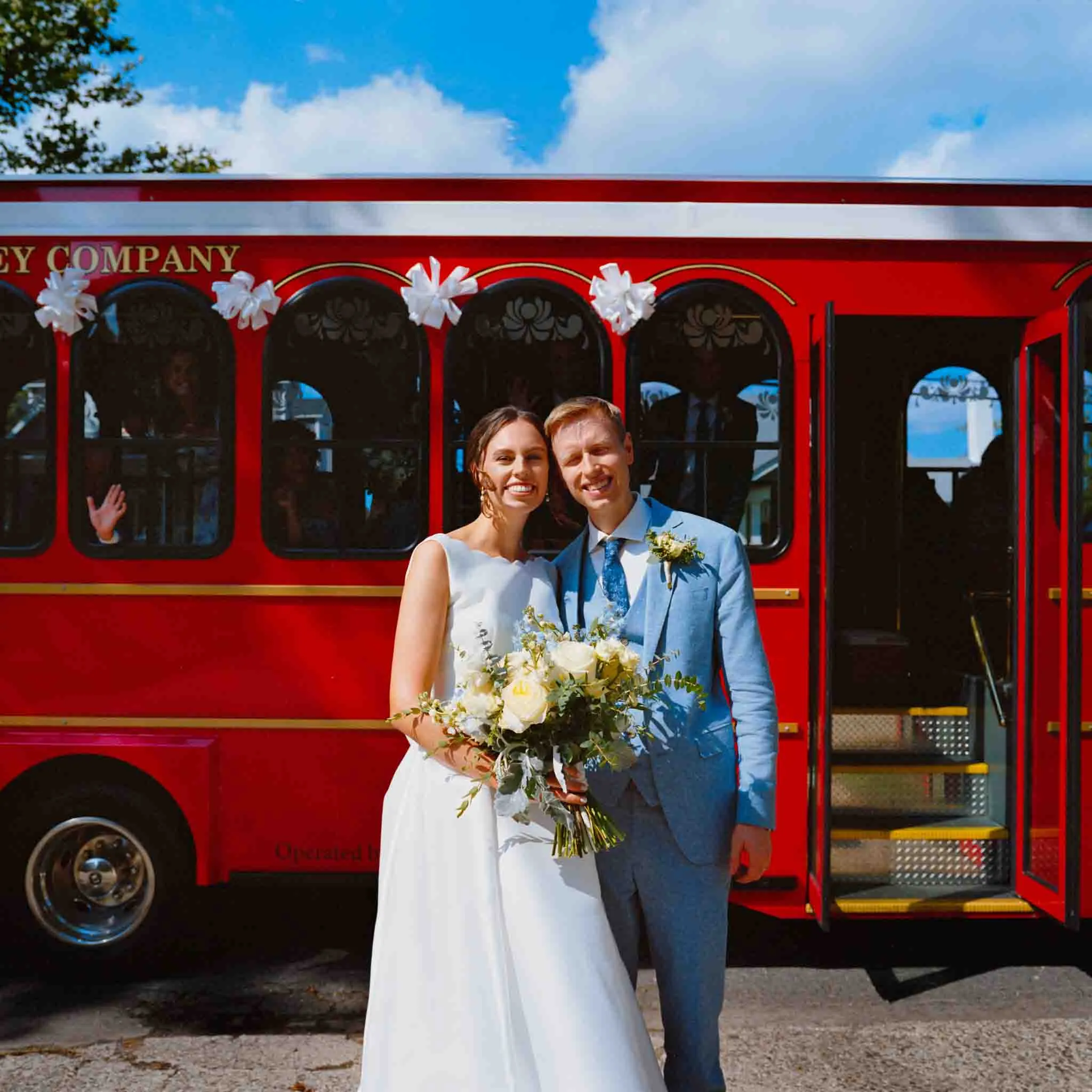 A bride and groom smile and pose together in front of a bright red trolley decorated with white bows on a sunny day. Captured by a documentary wedding photographer, the moment highlights the groom’s light blue suit and the bride’s bouquet.