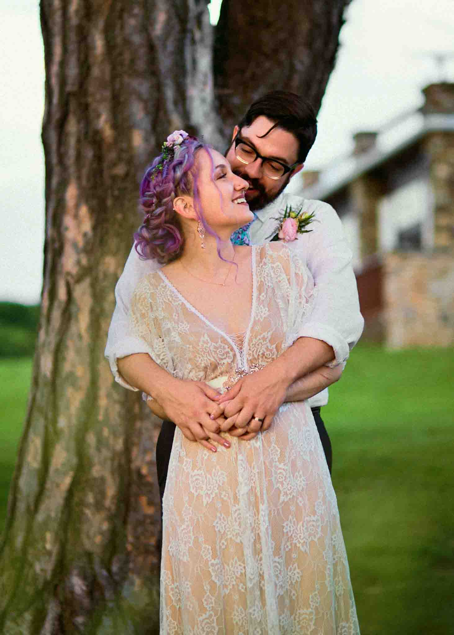 A couple stands outdoors by a tree, smiling and embracing. Captured by a film wedding photographer NJ, the woman in a lace dress with purple hair and the man with glasses look happy in a natural, grassy setting.
