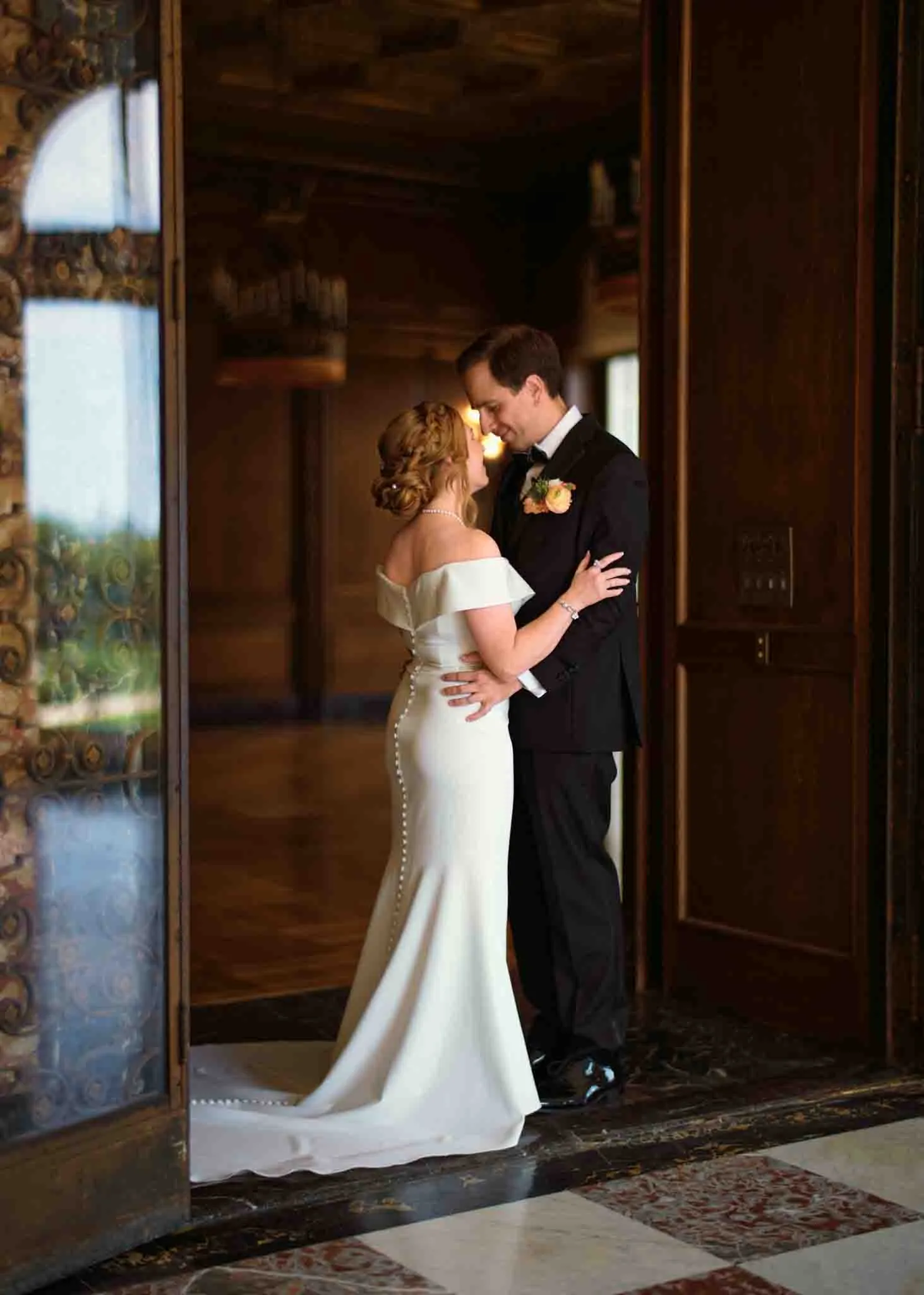 A bride in an off-shoulder, buttoned white gown and a groom in a black suit embrace and gaze at each other lovingly inside an elegant, warmly lit room—an intimate moment captured by a documentary wedding photographer.