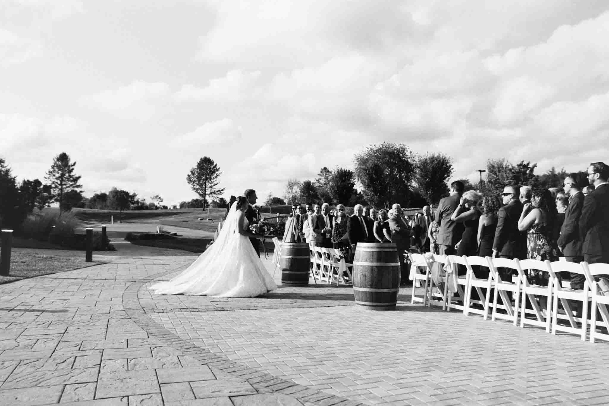 A bride in a flowing gown walks down an outdoor aisle lined with white chairs, as guests watch her. Captured by a New Jersey film wedding photography expert, the candid moment unfolds beneath trees and a partly cloudy sky.