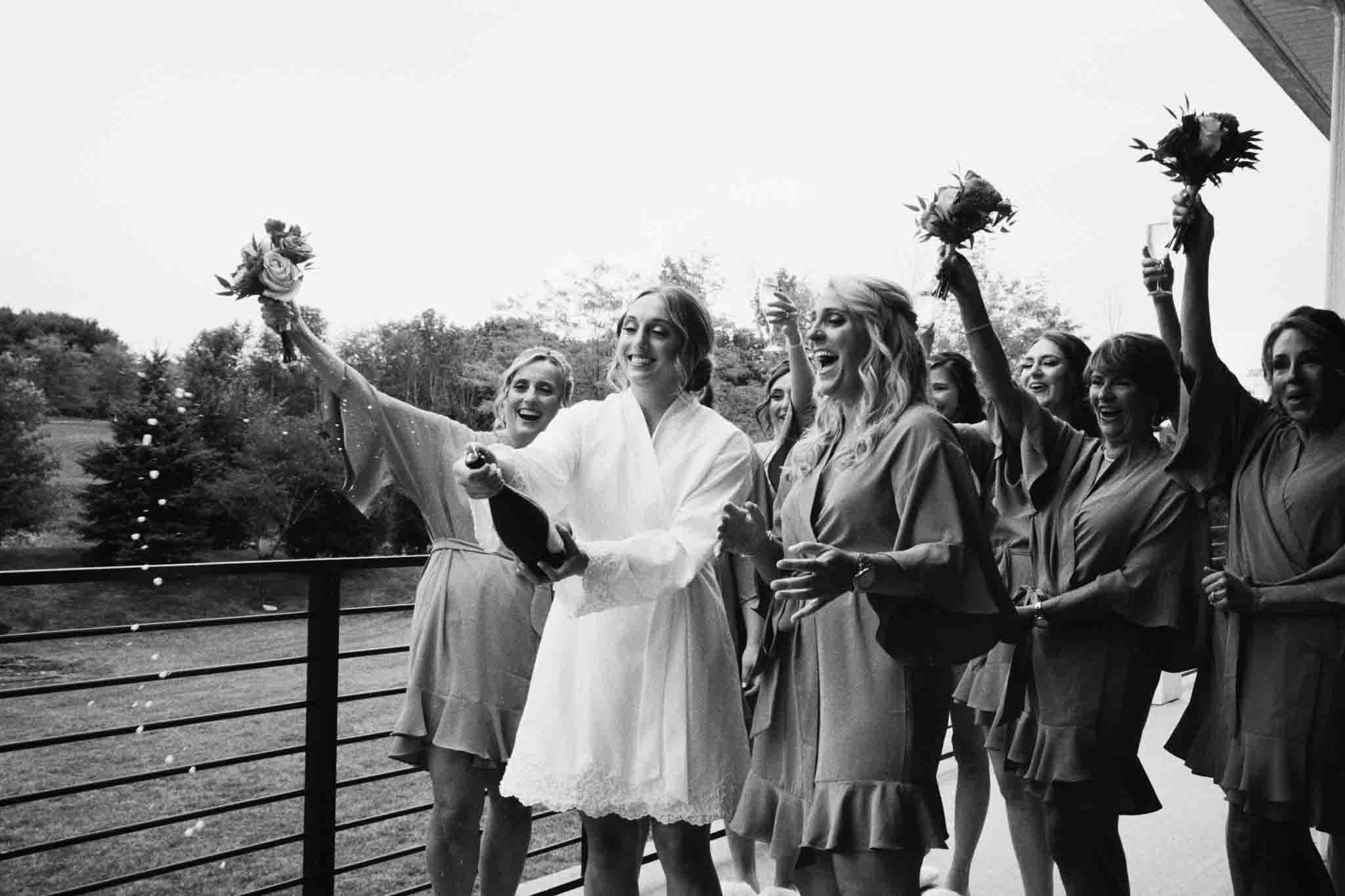 A bride in a white robe pops champagne, surrounded by bridesmaids in matching robes holding bouquets and cheering on a balcony. Captured in the candid film wedding photos style by a New Jersey film wedding photography expert.