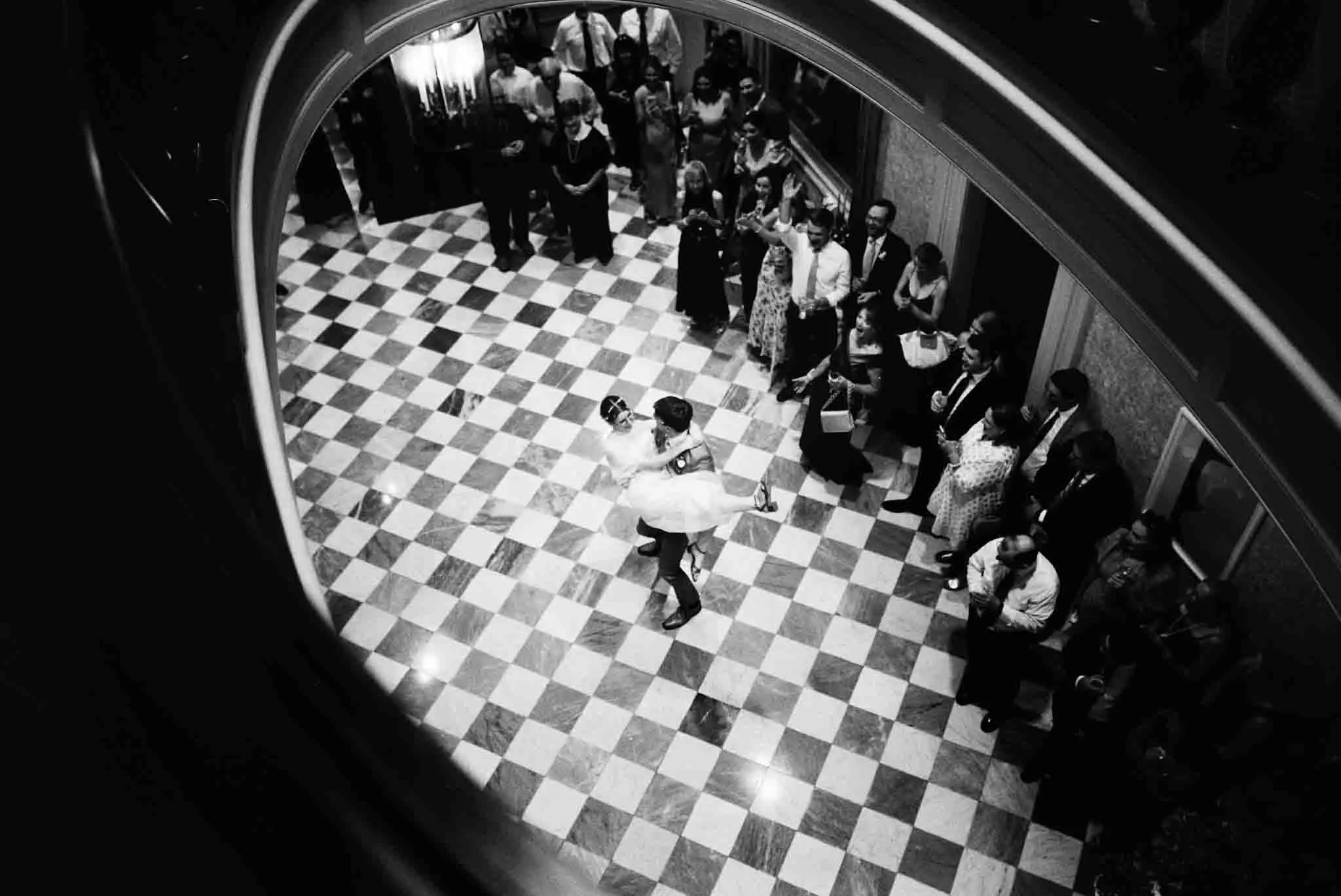 A bride and groom dance on a checkered floor as guests look on in an elegant venue, captured from above by a New Jersey black and white film wedding photographer.