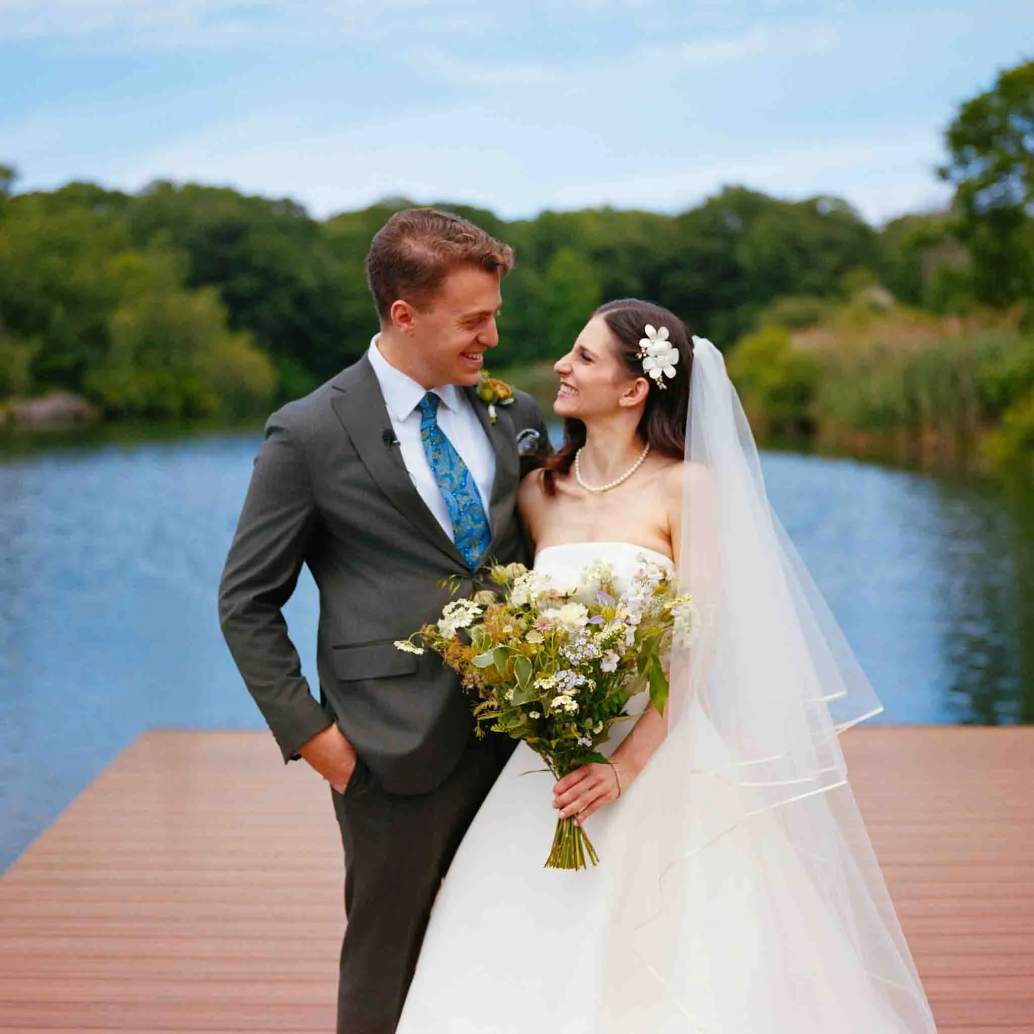 A bride and groom stand on a wooden dock by a lake, smiling at each other. Captured in the candid film wedding photos style by a documentary wedding photographer, the lush greenery and joyful moment reflect true connection in New Jersey.