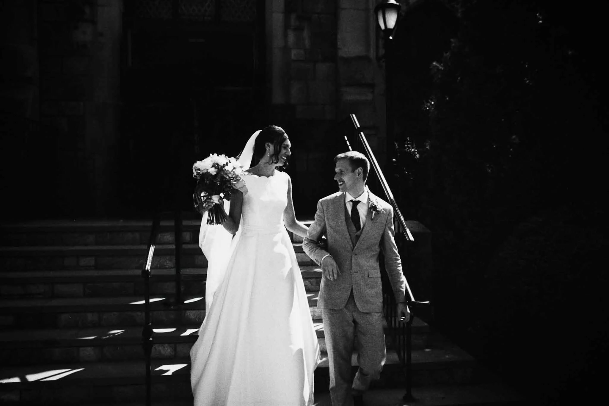 A bride in a white dress and veil walks arm-in-arm with a groom in a light suit, smiling at each other as they descend stone steps, sunlight casting dramatic shadows—evoking candid film wedding photos New Jersey couples cherish.