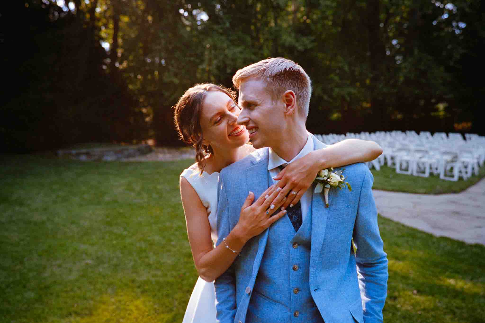 A smiling bride in a white dress hugs a groom in a light blue suit from behind. Captured with candid film wedding photos New Jersey style, they stand outdoors on grass, with white chairs and trees in the background, bathed in warm sunlight.