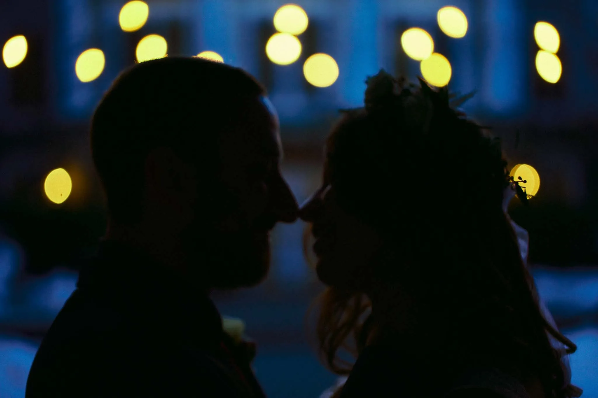 A silhouetted couple faces each other closely, possibly about to kiss, with blurred warm lights glowing in the background and a soft blue evening ambiance—a moment beautifully captured by a film wedding photographer NJ.