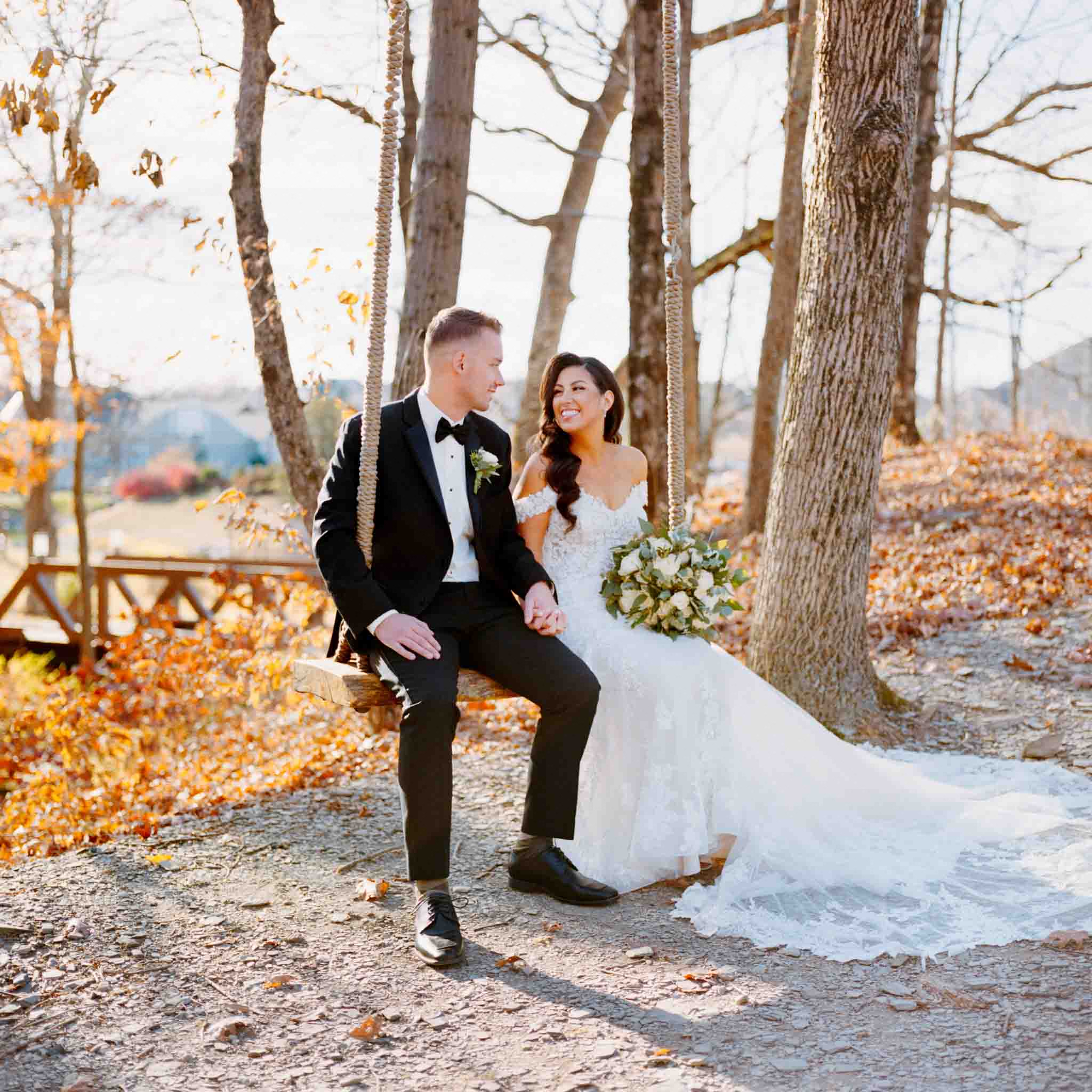 A bride in a white gown and a groom in a black suit sit together on a wooden swing amid autumn leaves, smiling and holding hands, with a bouquet in the bride’s lap—captured in the timeless style of candid film wedding photos New Jersey.
