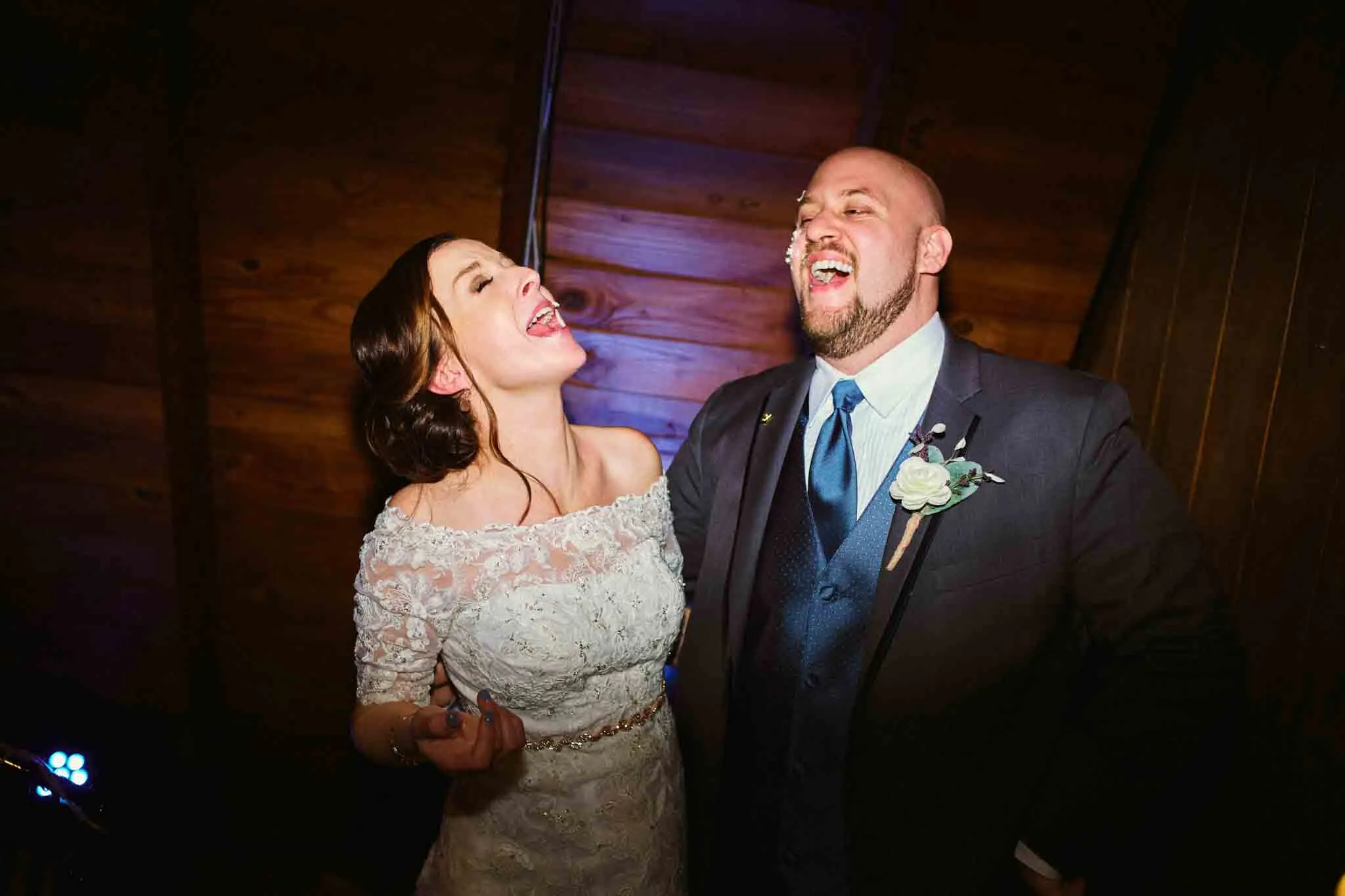 A bride and groom in wedding attire share a joyful laugh, standing close under warm indoor lighting with a wooden ceiling in the background—a candid moment captured by a documentary wedding photographer specializing in analog wedding photography NJ.