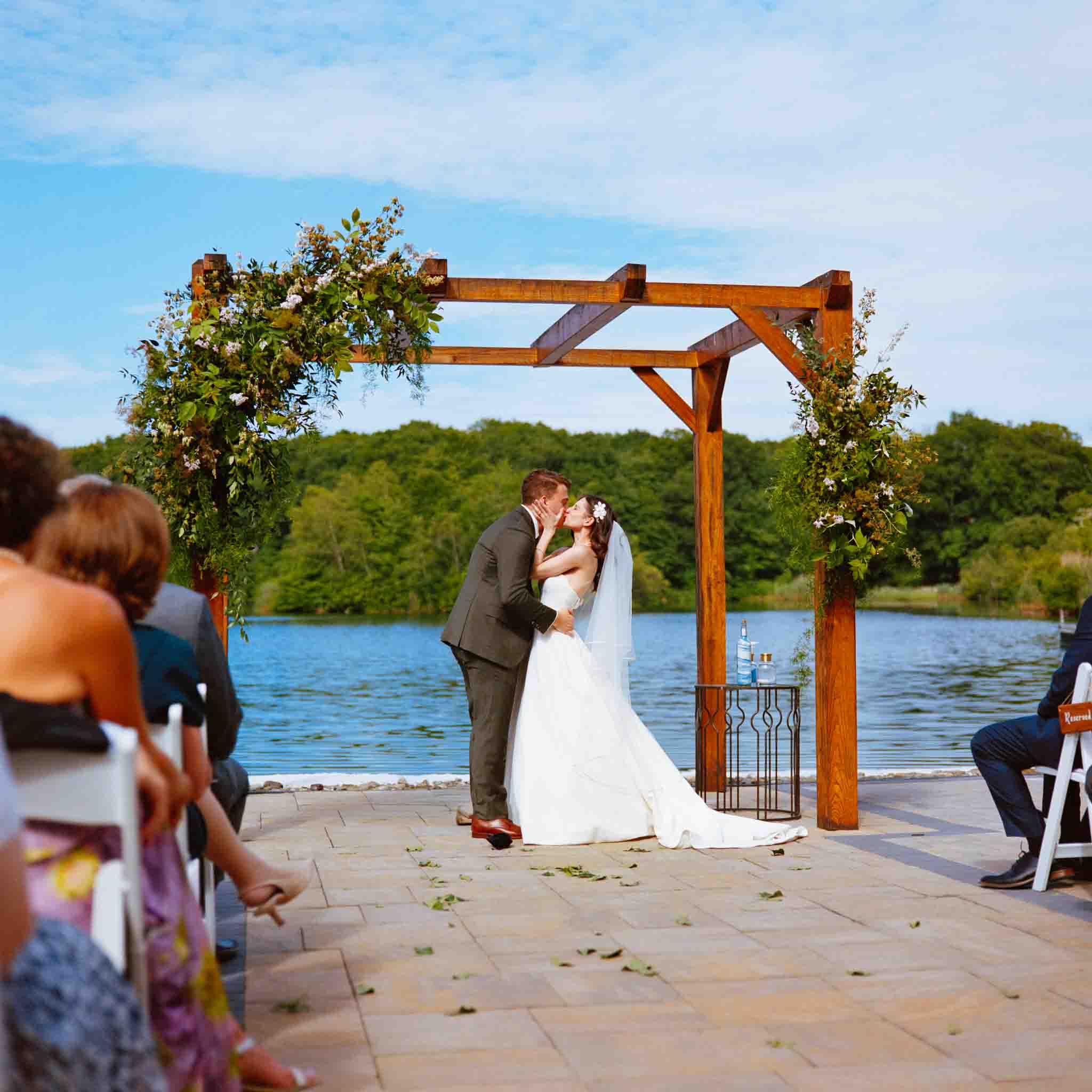 A bride and groom kiss under a wooden arch adorned with greenery during an outdoor lakeside wedding, beautifully captured by a wedding photographer specializing in candid film wedding photos New Jersey. Guests are seated on either side.