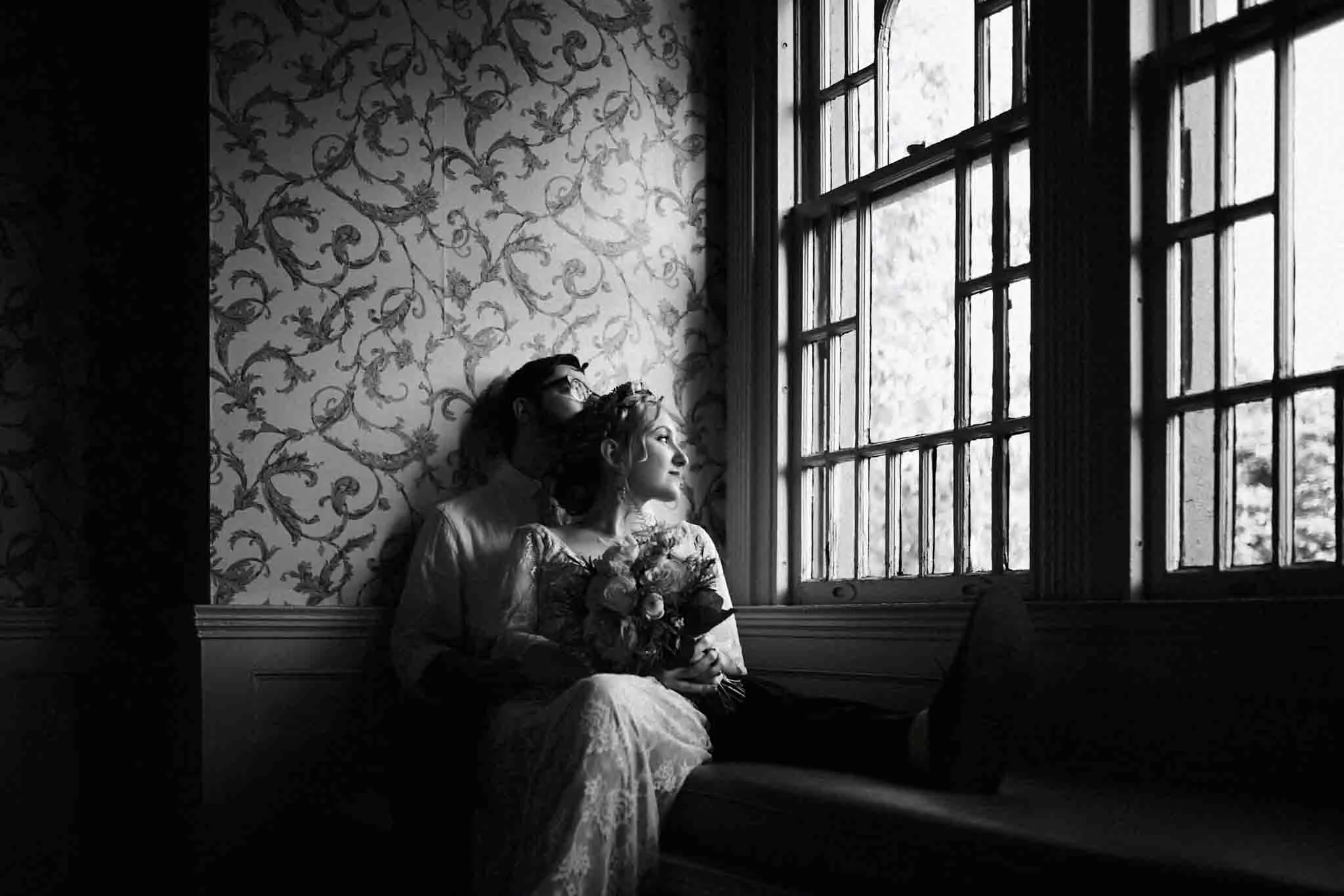 A bride and groom sit closely on a window seat, gazing outside. The bride holds a bouquet in her lace dress as soft light filters through large windows in a vintage, patterned room. Captured by a New Jersey film wedding photographer in black and white.