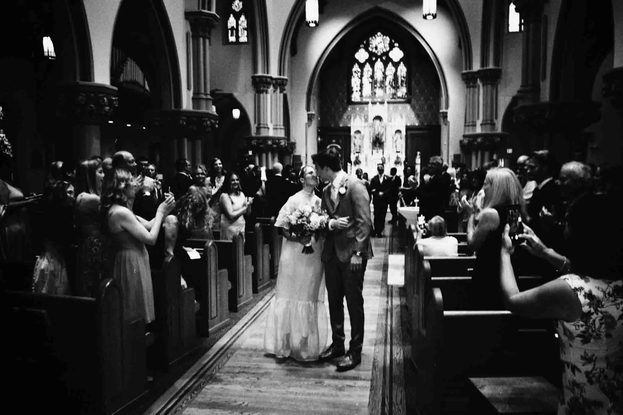 A newlywed couple walks down the aisle of an ornate church as guests stand and take photos. The setting, perfect for a film wedding photographer NJ, features high ceilings and stained glass, creating a joyful and celebratory atmosphere.
