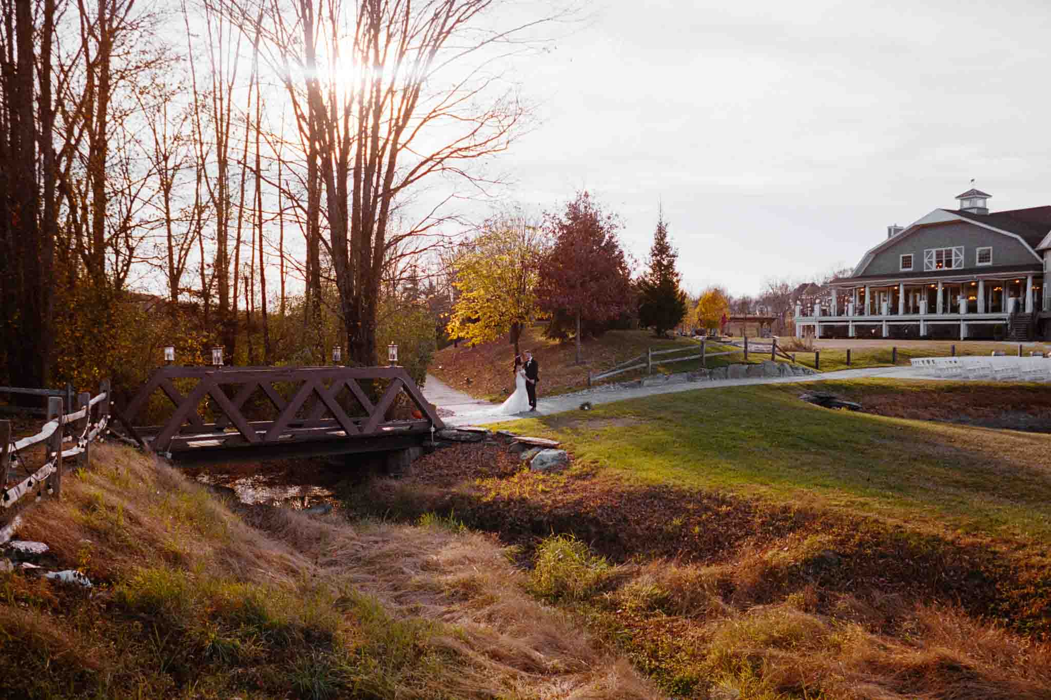 A couple walks hand in hand across a small wooden bridge in a sunlit, autumn landscape, captured by an analog wedding photographer. Trees, grassy hills, and a large house with a porch create a picturesque New Jersey backdrop.