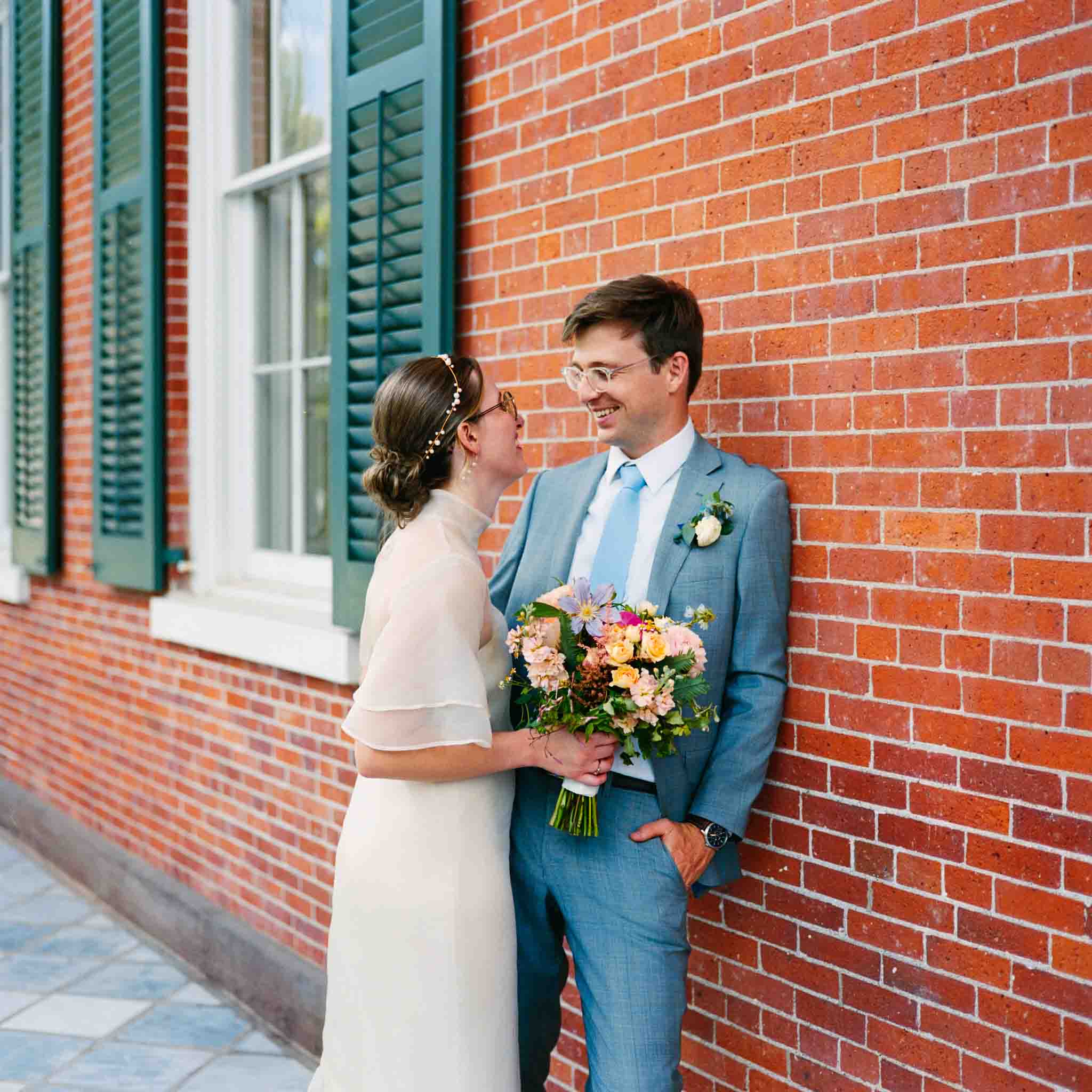 A bride in a white dress and a groom in a blue suit smile at each other against a red brick wall with green shutters. The bride holds a colorful bouquet, captured by an analog wedding photography NJ specialist.