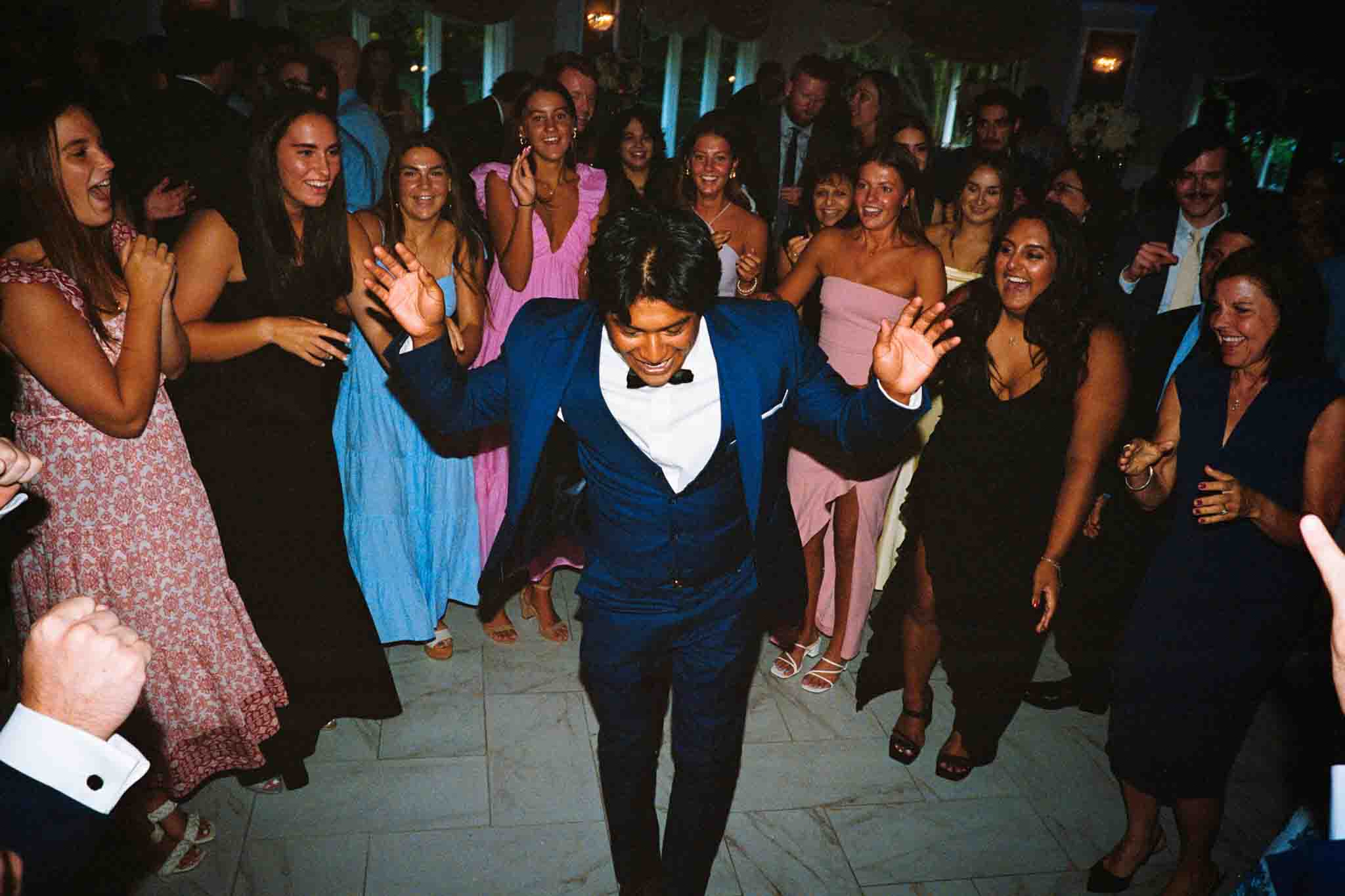 A man in a blue suit dances at the center of a lively group at an indoor wedding reception. Surrounded by smiling guests in formal attire, he is captured with excitement and joy by a documentary wedding photographer.