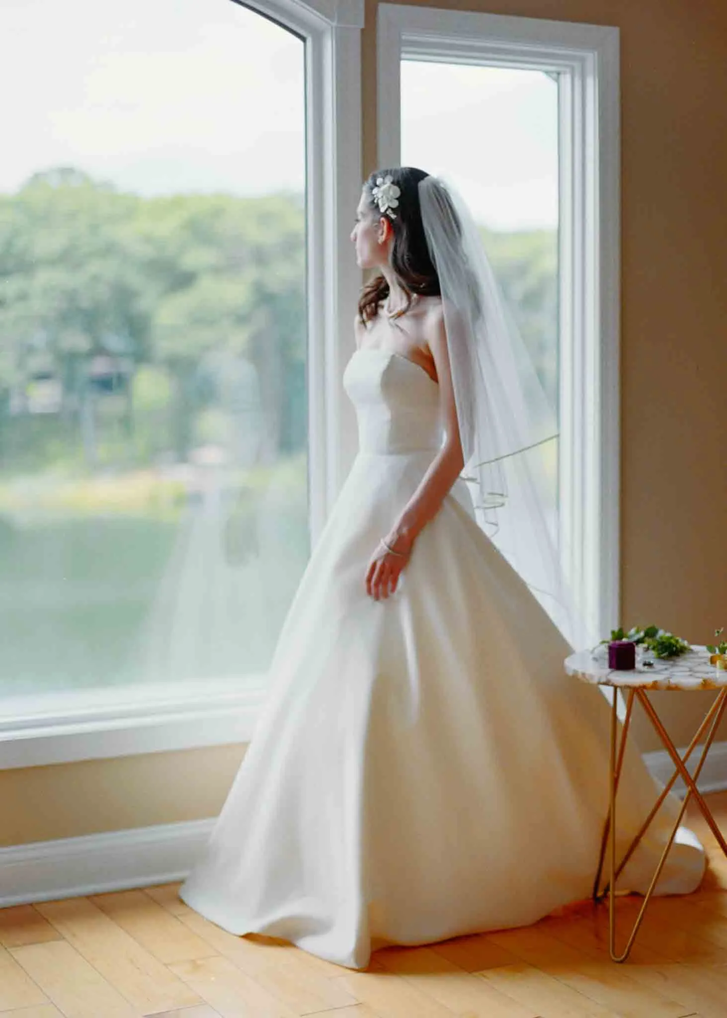 A bride in a white strapless gown and veil stands by a large window, gazing outside at a green landscape. Captured with the artistry of New Jersey film wedding photography, flowers and a ring box rest on a small table nearby.