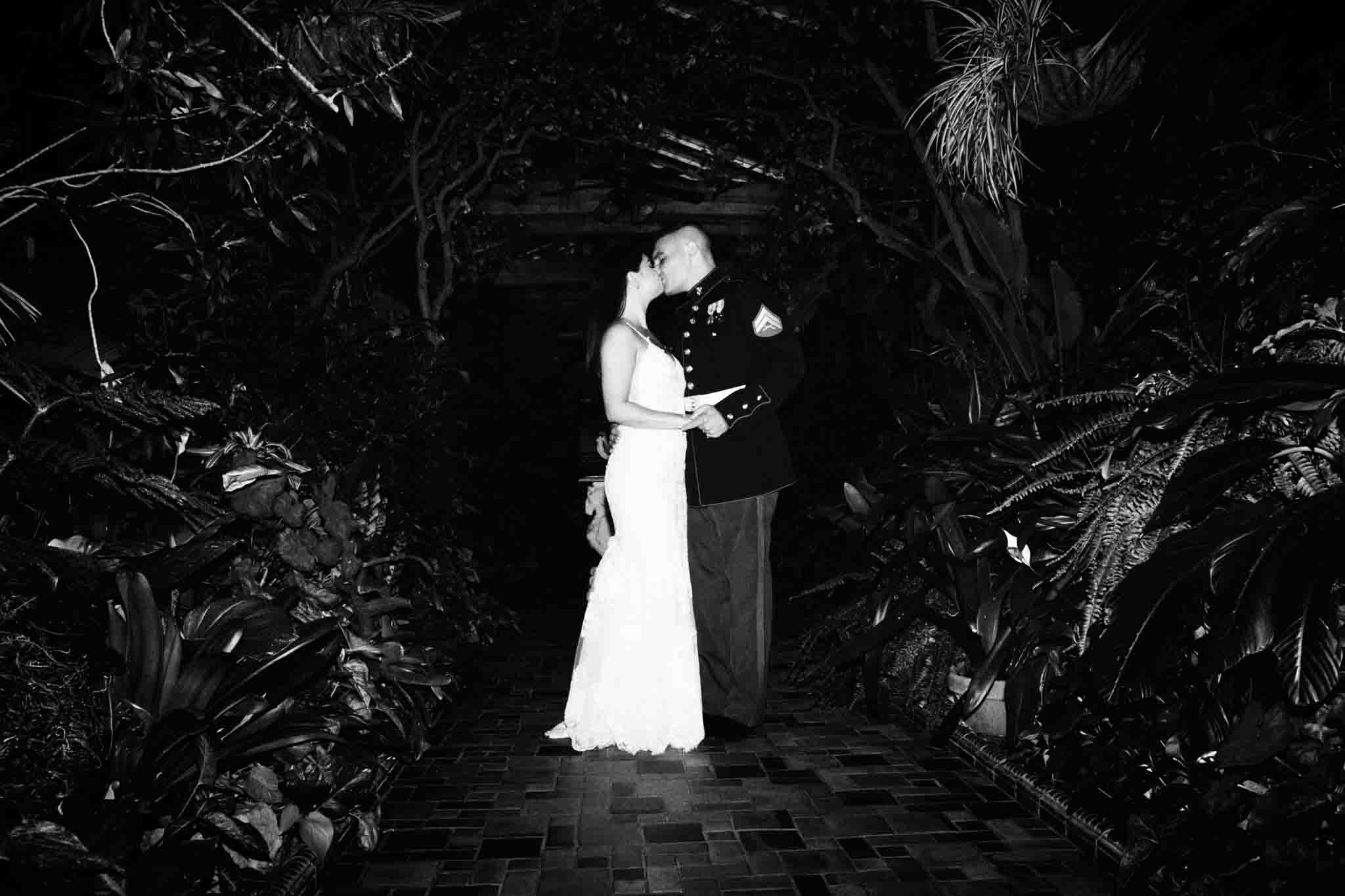 A couple kisses in the center of a lush, leafy garden at night. The woman wears a white dress and the man a formal military uniform—an enchanting scene ideal for candid film wedding photos New Jersey couples cherish.