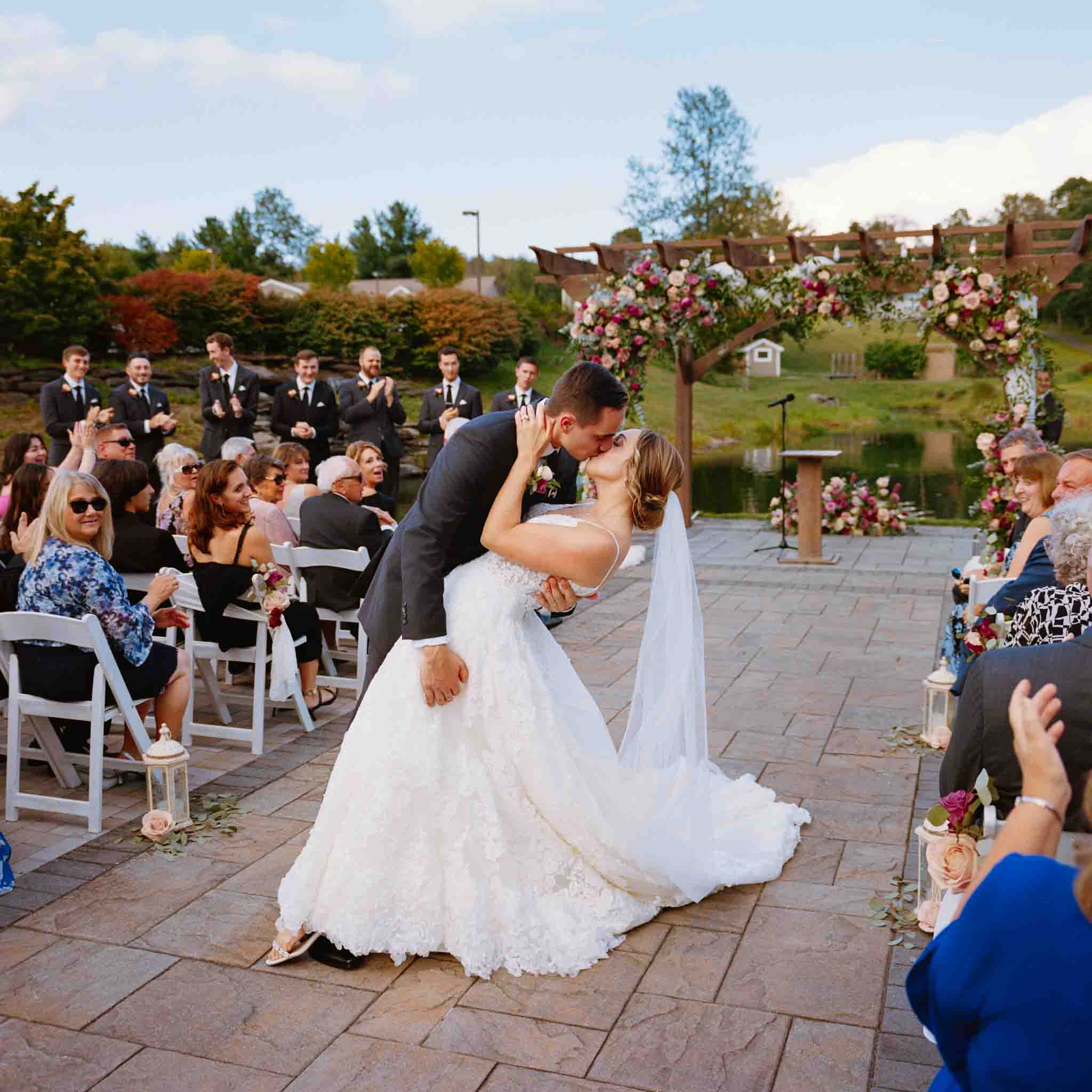 A bride and groom share a kiss at an outdoor wedding ceremony, surrounded by guests clapping and smiling. Floral arrangements and a wooden arch frame the scene, captured beautifully by a wedding photographer specializing in candid film wedding photos New Jersey.