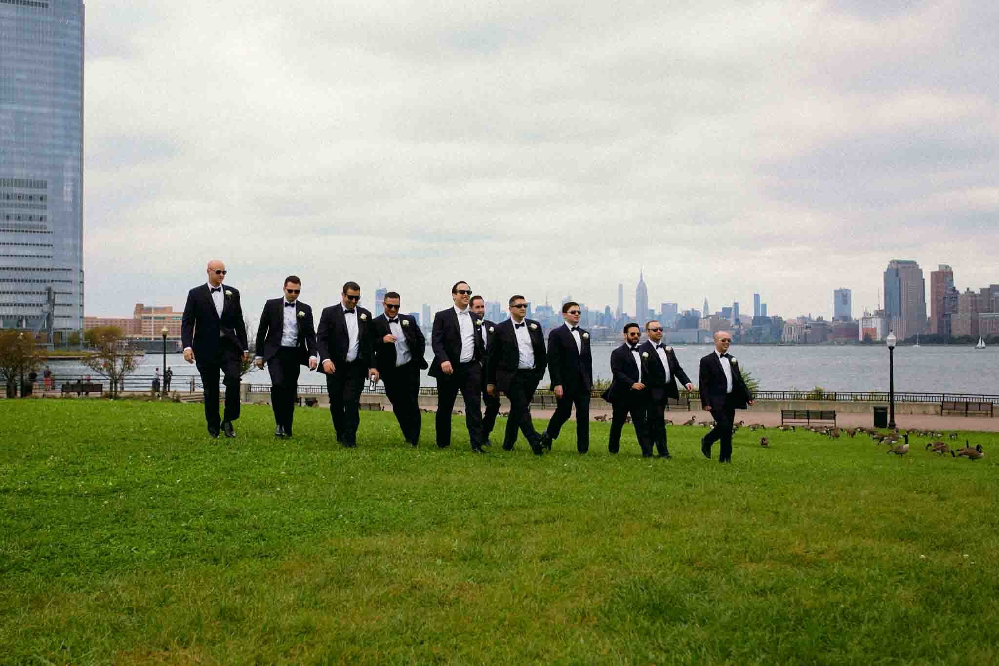 A group of men in black tuxedos and sunglasses walk confidently across a grassy park, with a city skyline and river in the background on a cloudy day—perfect for candid film wedding photos New Jersey style.