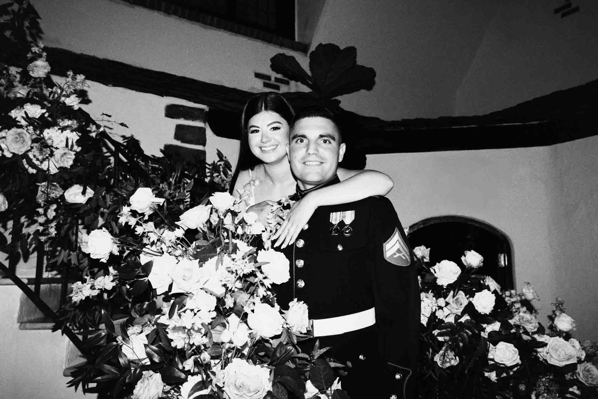 A smiling woman embraces a man in military dress uniform as they stand amid abundant flower arrangements on a staircase, both looking at the camera in this black-and-white candid film wedding photo by a New Jersey film wedding photographer.