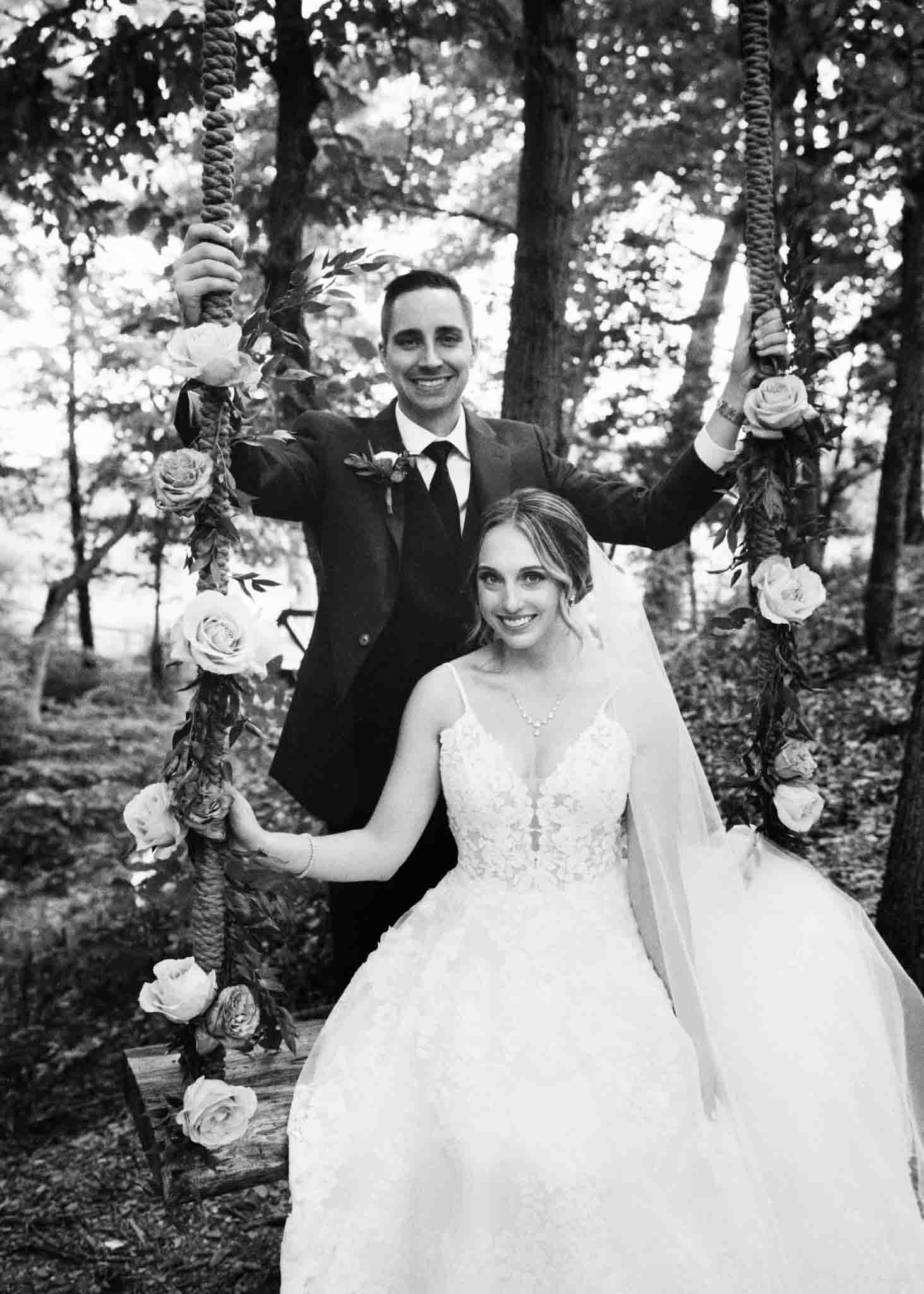 A bride in a lace wedding dress sits on a flower-decorated swing, smiling, while the groom in a suit stands behind her in a wooded outdoor setting—captured beautifully by a New Jersey film wedding photographer.