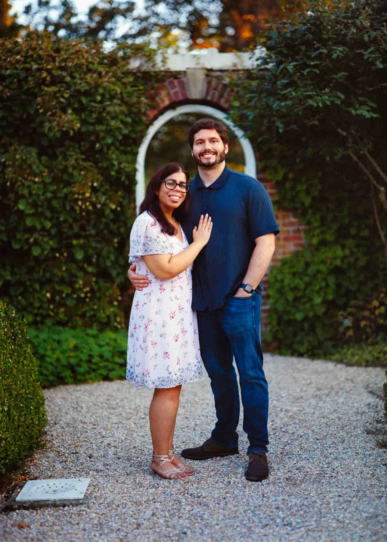 A smiling couple stands together on a gravel path in a garden, with greenery and a brick archway behind them. Captured by a documentary wedding photographer, they radiate joy—she in a white floral dress and glasses, he in a navy shirt and jeans.
