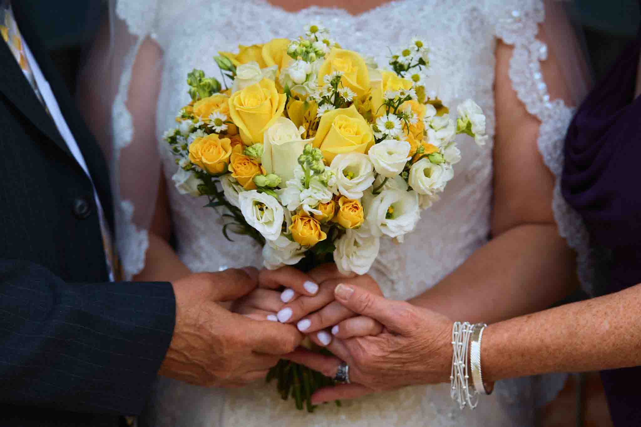 A bride in a white lace dress holds a bouquet of yellow and white flowers, with two people supportively touching her hands. Captured by a documentary wedding photographer, only their hands and part of their outfits are visible.