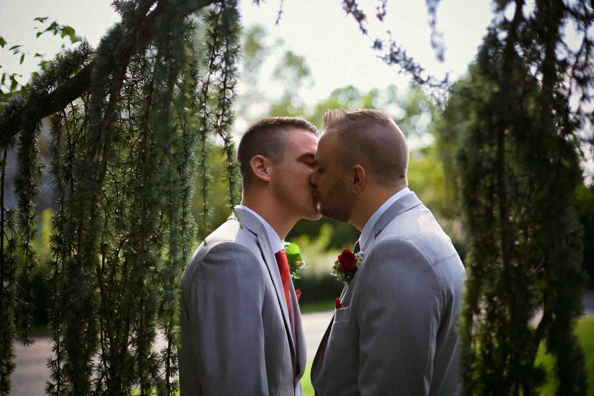 Two men in light gray suits with red boutonnieres share a kiss outdoors, framed by hanging tree branches and greenery—captured in authentic New Jersey film wedding photography style.