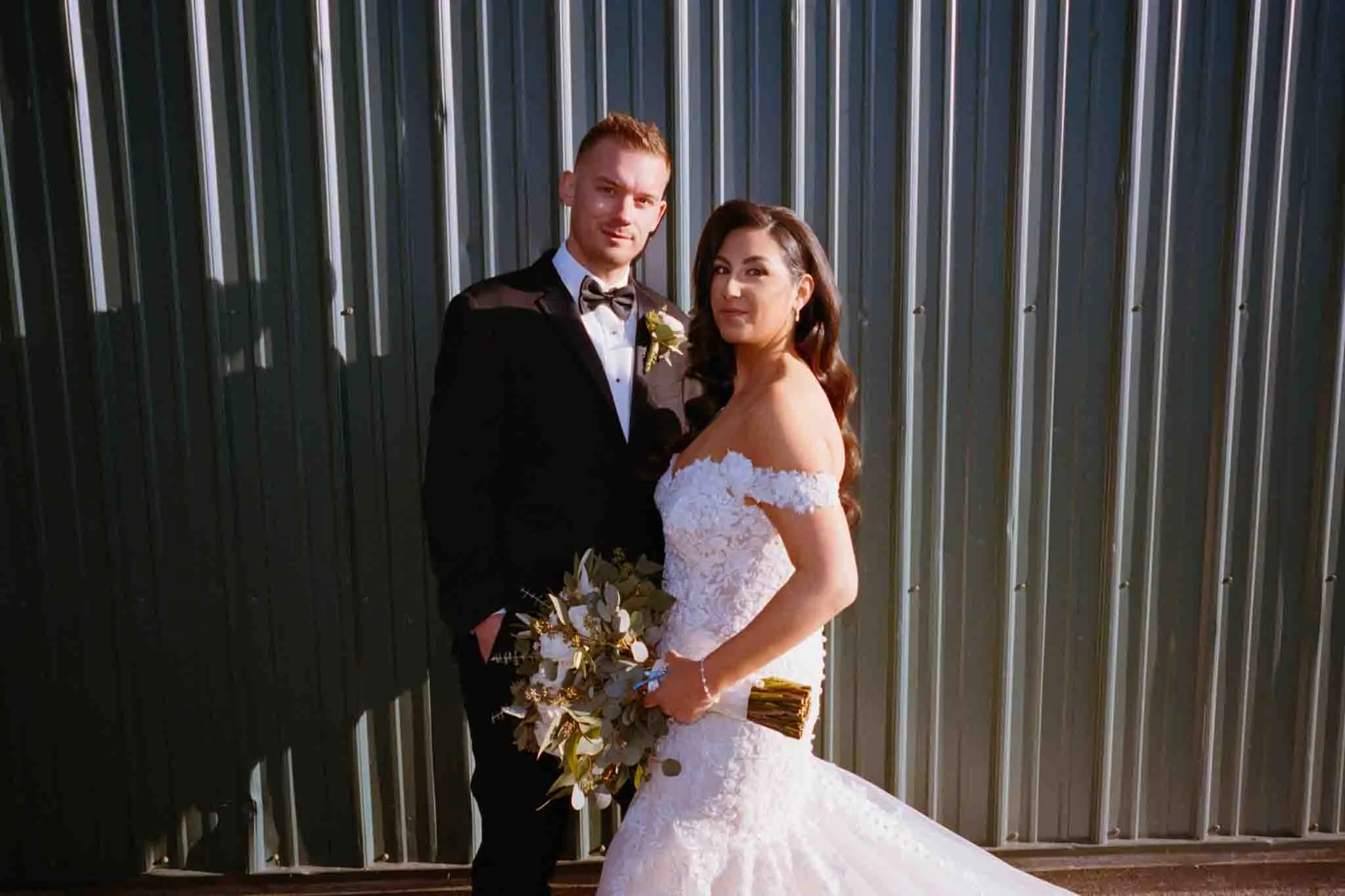 A bride in an off-shoulder white gown holds a bouquet and stands with her groom in a black tuxedo. Captured by a film wedding photographer NJ, they pose together before a corrugated metal wall, looking at the camera.