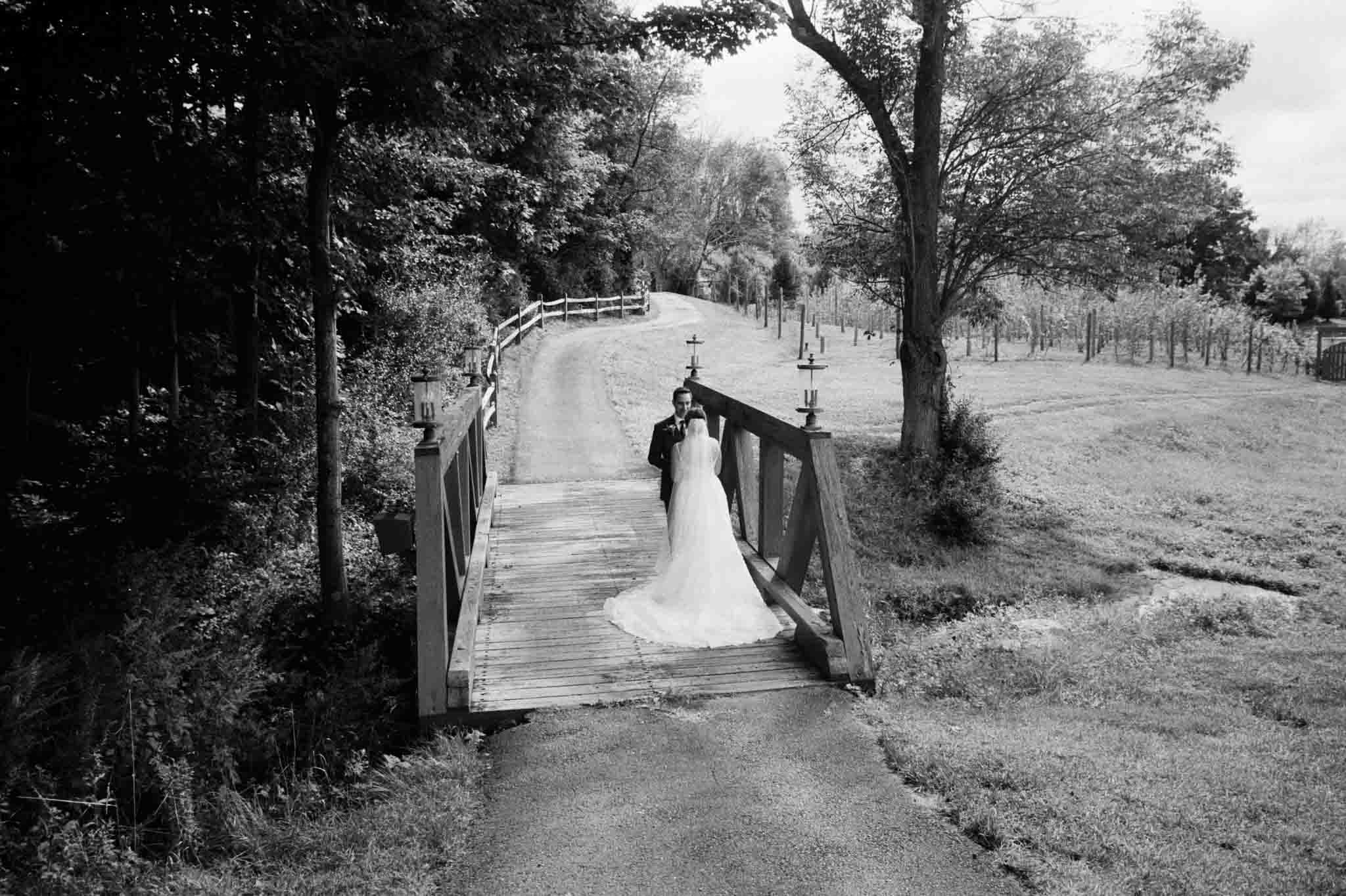 A bride and groom stand closely on a small wooden bridge surrounded by trees and fields. Captured by a documentary wedding photographer in black and white, the scene is peaceful, timeless, and full of romance.