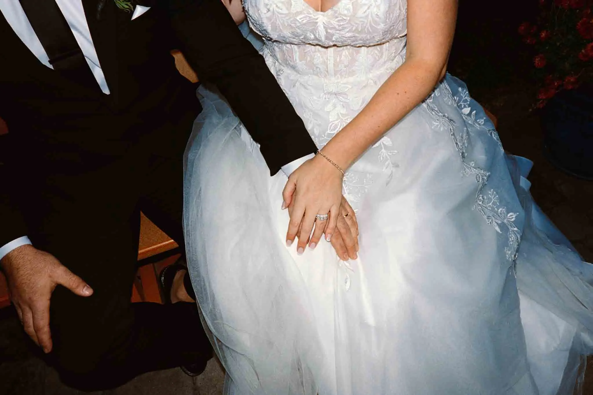 A bride in a white lace wedding dress sits closely with her groom in a black suit, her wedding ring sparkling as their hands rest together. Captured by a New Jersey film wedding photographer, this moment radiates love and elegance.
