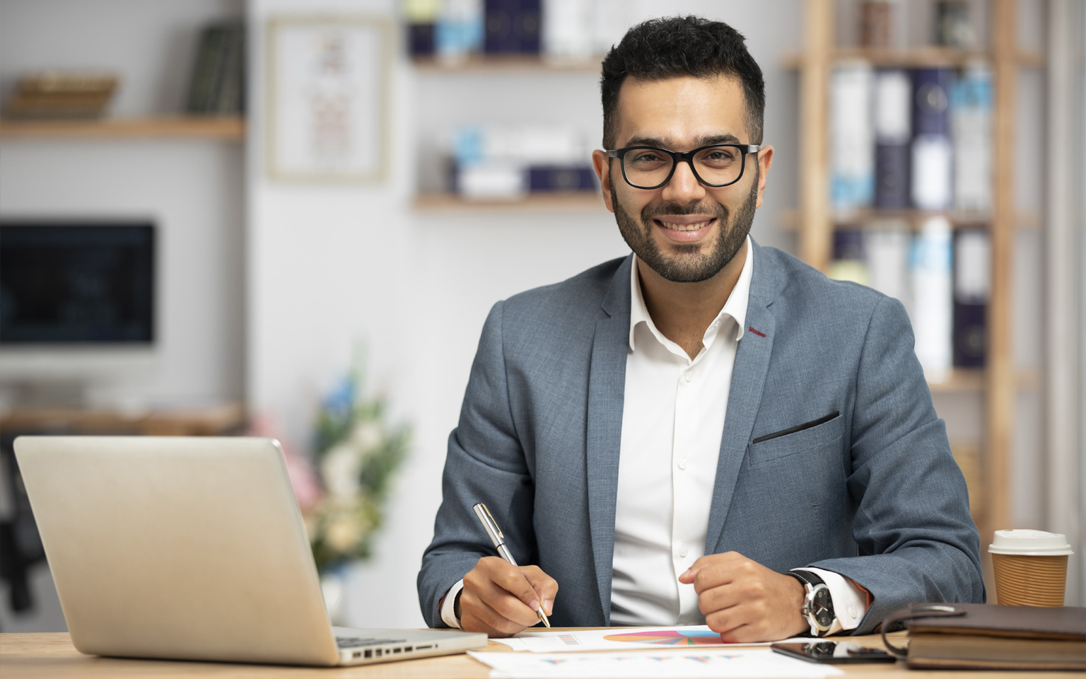 Smiling man with glasses in a suit sitting at a desk with a laptop, papers showing charts, and a coffee cup.