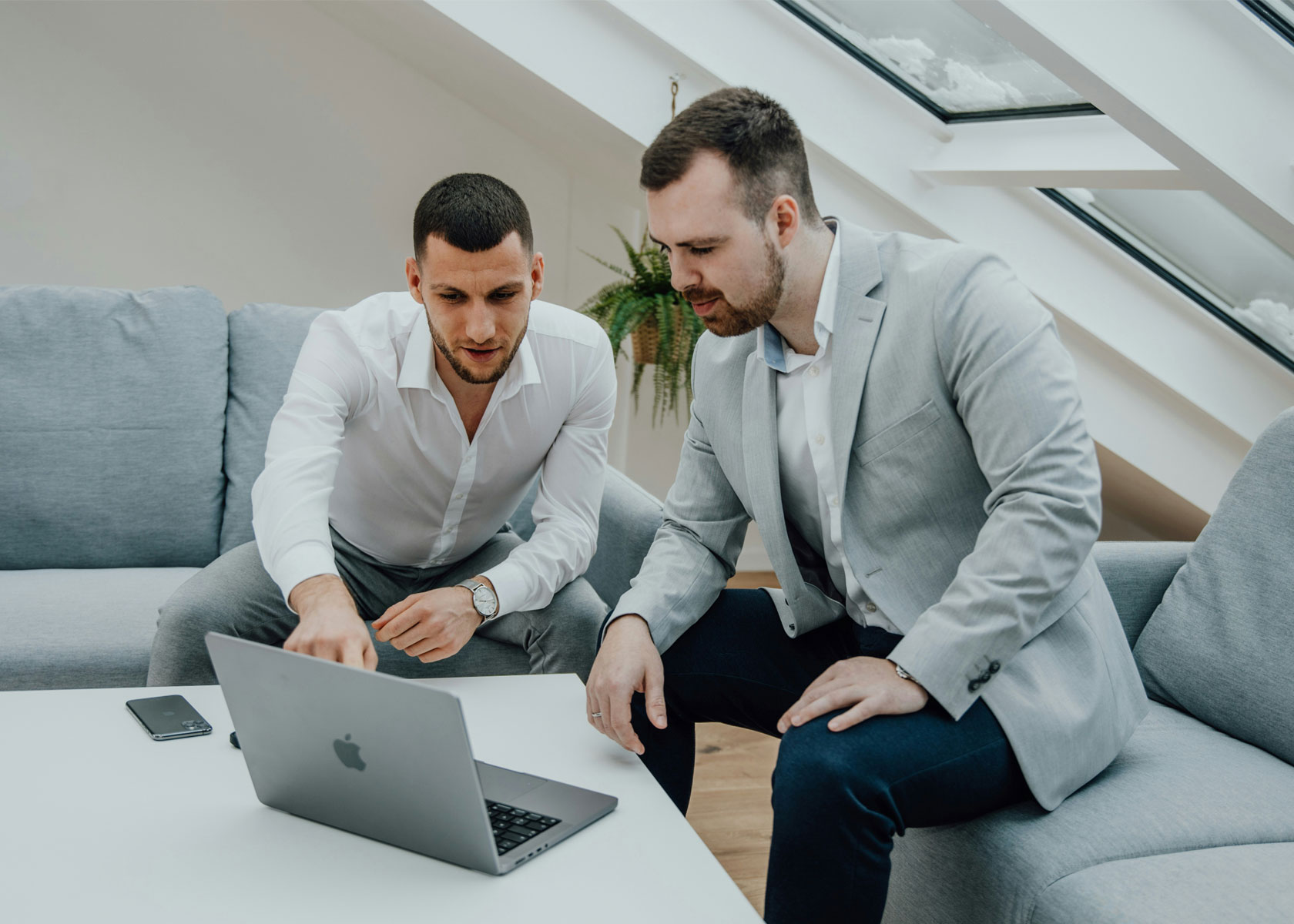 Two men, one in a white shirt and one in a light gray blazer, sitting on a gray sofa and looking at a laptop on a white table.