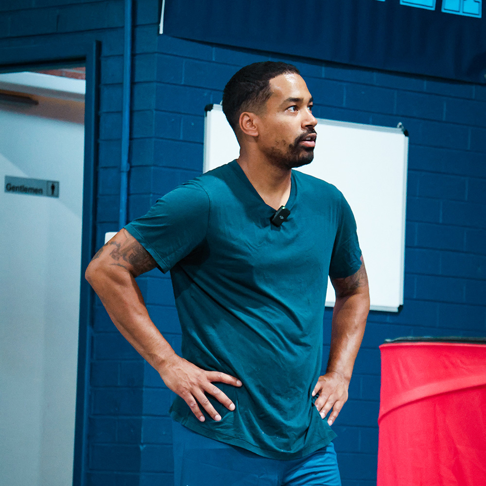 Man with short curly hair, wearing a teal T-shirt, standing with hands on hips in an indoor gym or sports facility.