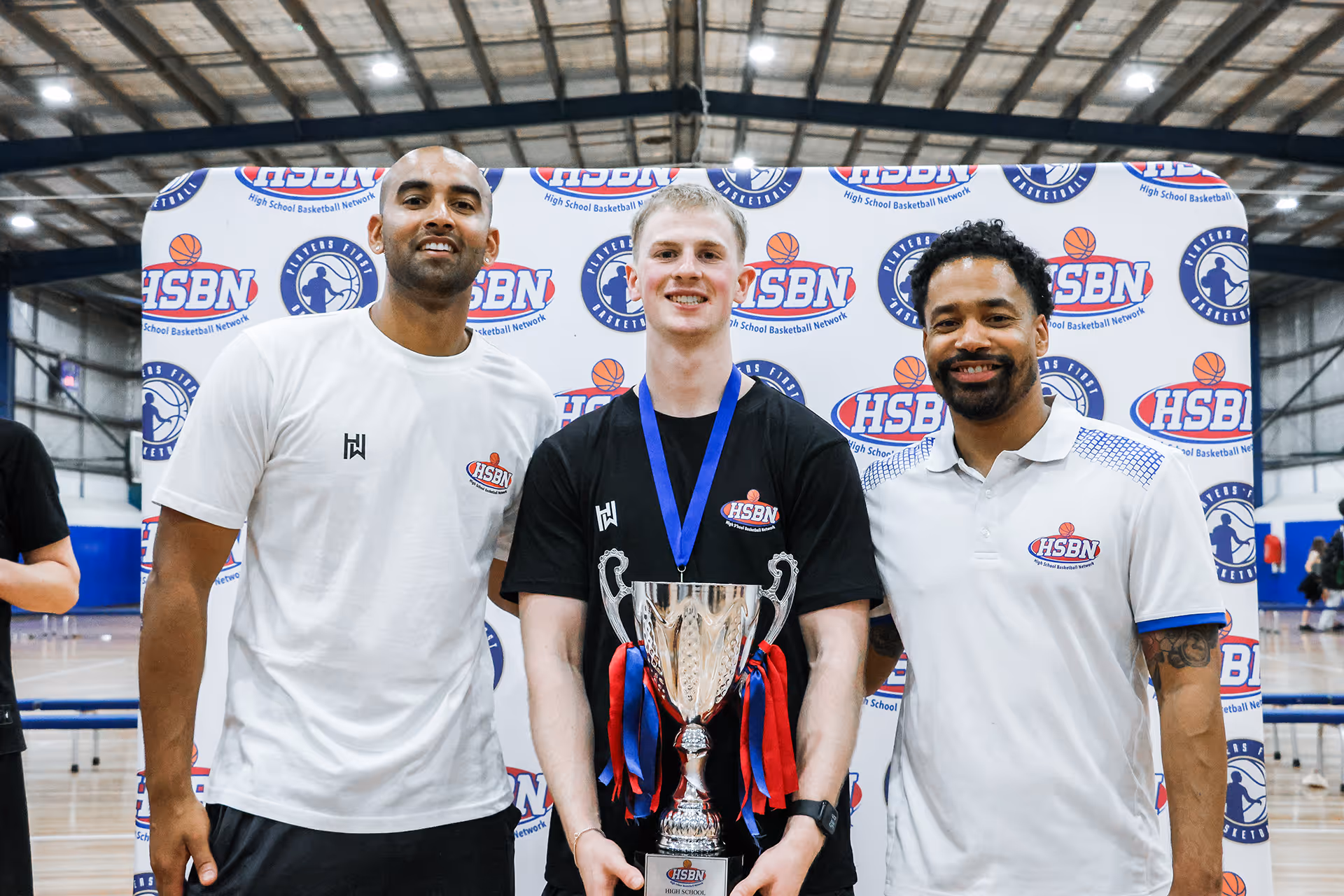 Three men standing in front of a High School Basketball Network banner, the man in the center holding a large trophy with red and blue ribbons and wearing a medal.
