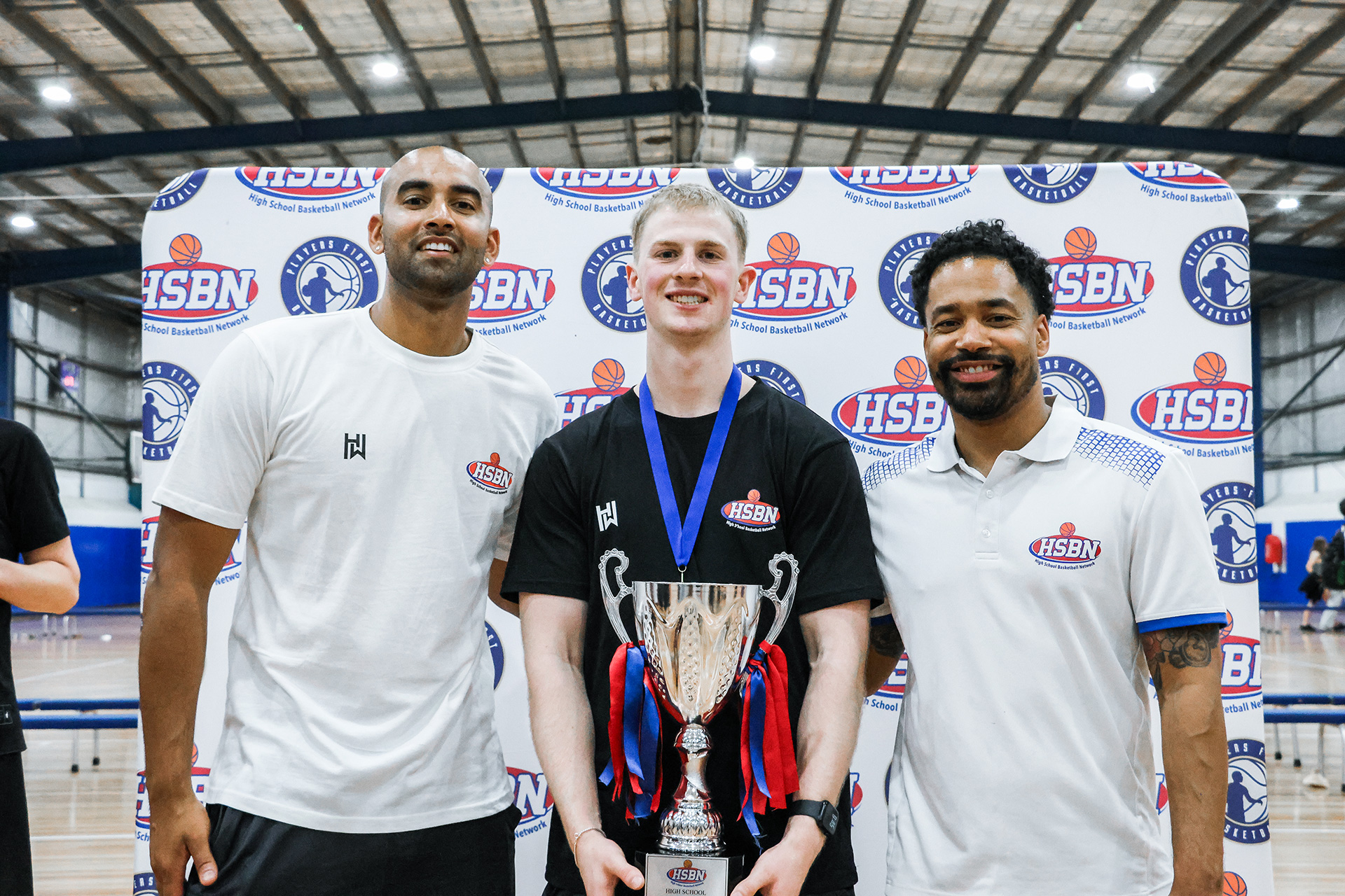 Three men standing in front of a High School Basketball Network banner, the man in the center holding a large trophy with red and blue ribbons and wearing a medal.