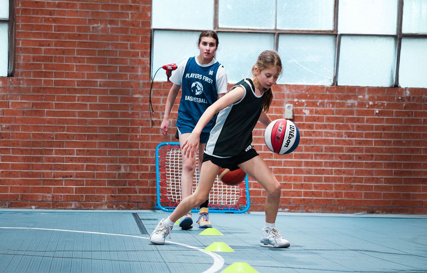 Two girls practicing basketball dribbling drills indoors on a blue court with a brick wall background.
