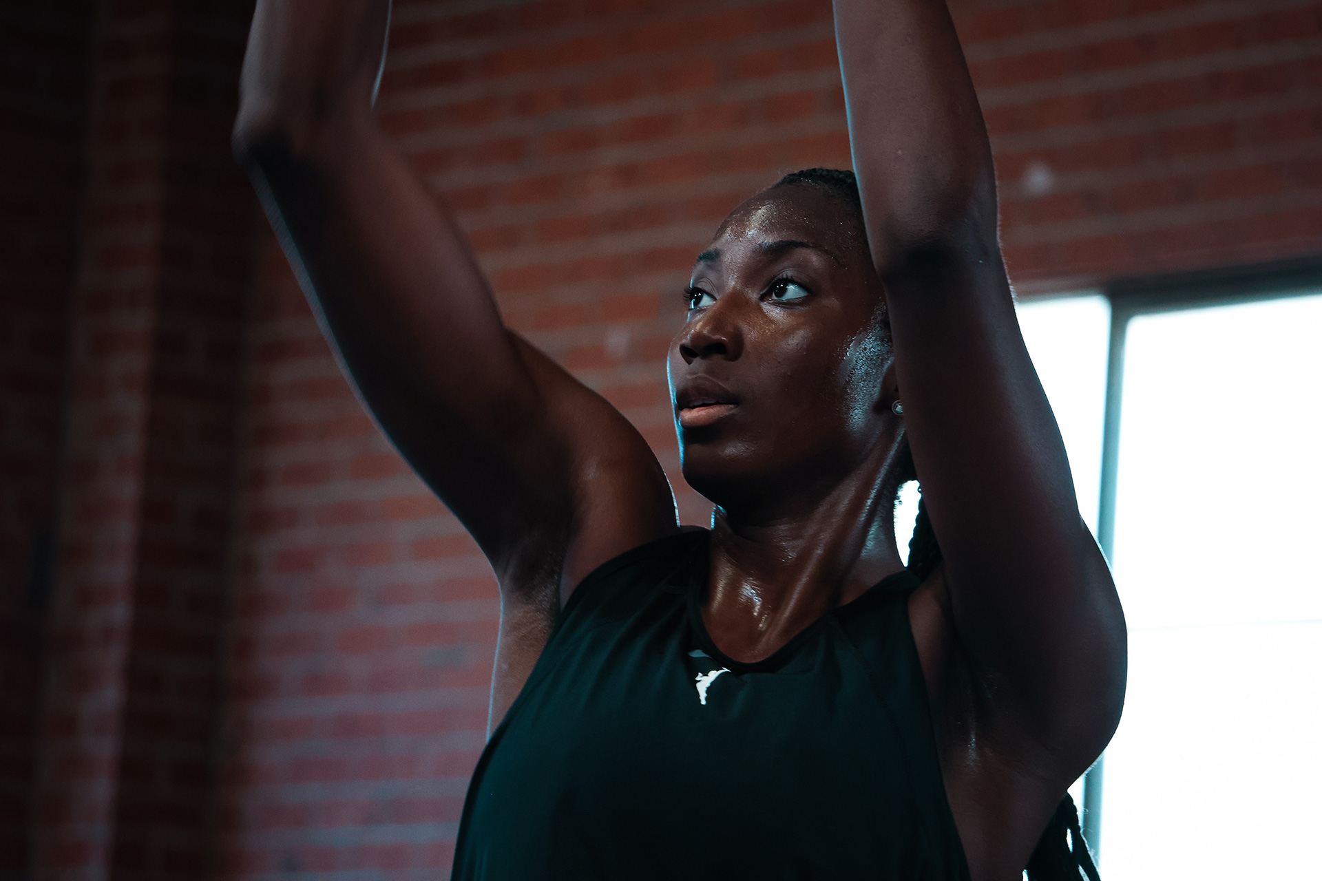 Focused young woman lifting arms during workout in a gym with brick wall background.