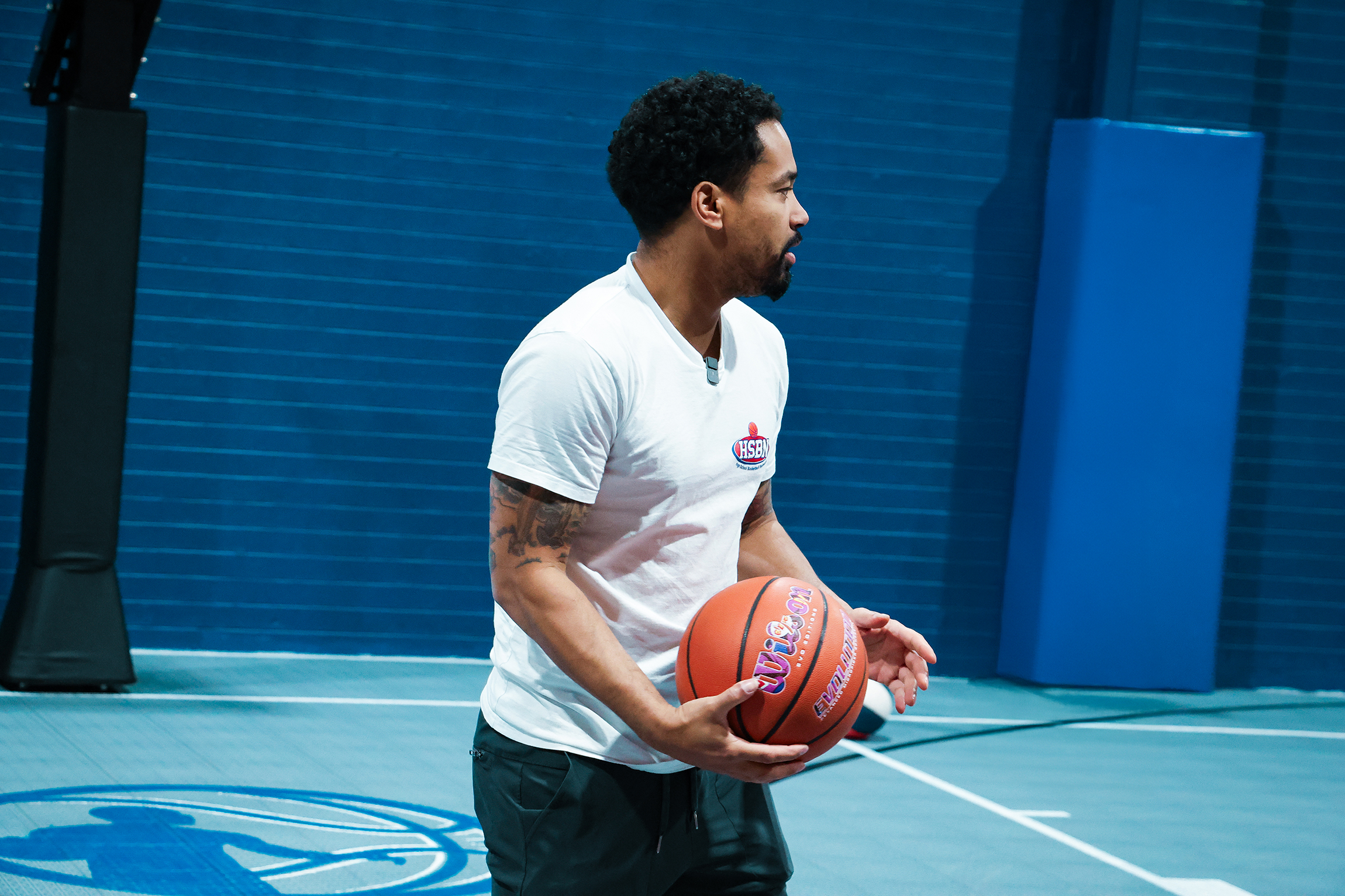Man in white t-shirt holding a basketball on an indoor court with blue walls and padded posts.