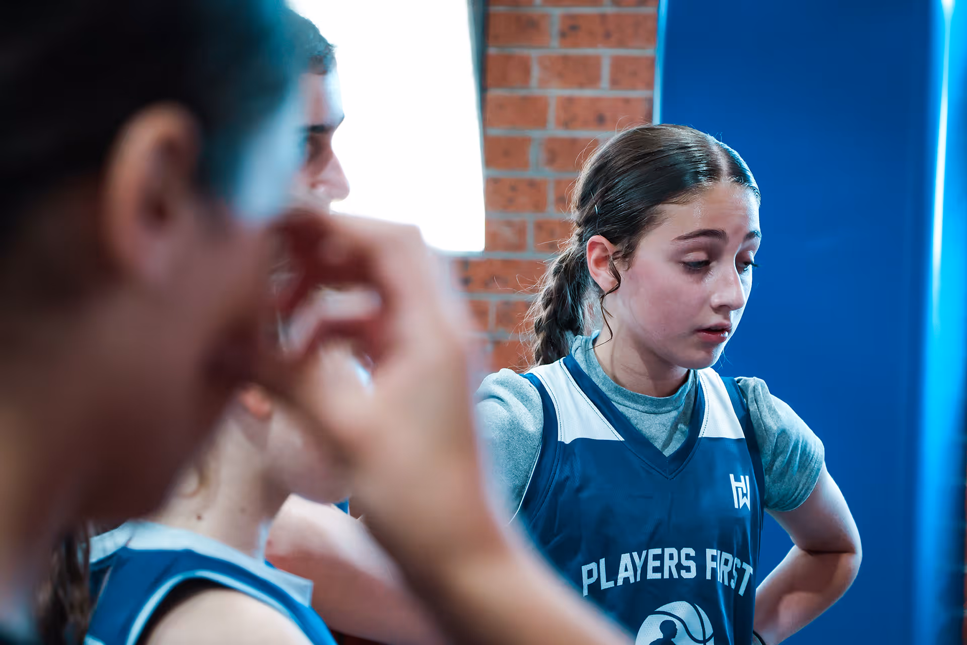 Young female basketball player with hands on hips, wearing a blue 'Players First' jersey, appears thoughtful during a break.