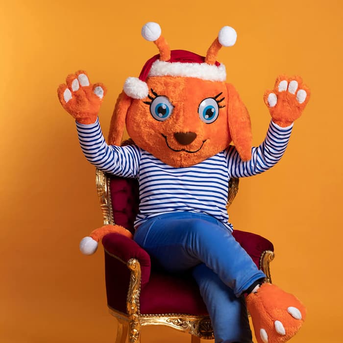 Maskottchen Luna im Weihnachtsoutfit sitzt fröhlich auf einem roten Sessel vor orangem Hintergrund