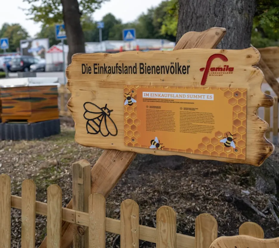 Holzschild mit der Aufschrift „Die Einkaufsland Bienenvölker“ auf dem Gelände von famila Einkaufsland Wechloy, Teil des Bienenprojekts für Umweltbildung.