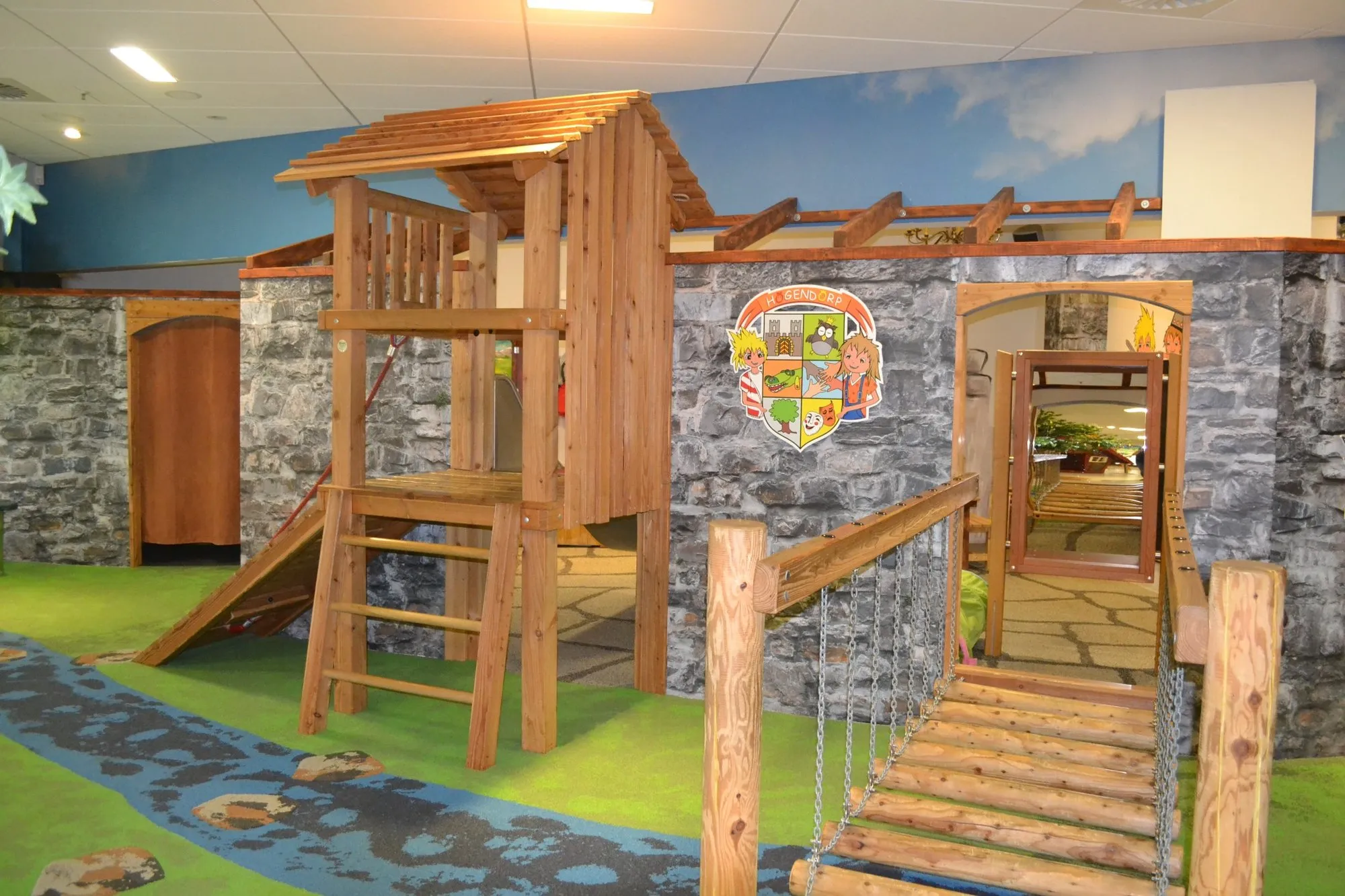 Holzspielhaus im „Högendörp“ mit Kletterelementen, daneben Öffnungszeiten und Altersangabe für den Kinderbereich.   