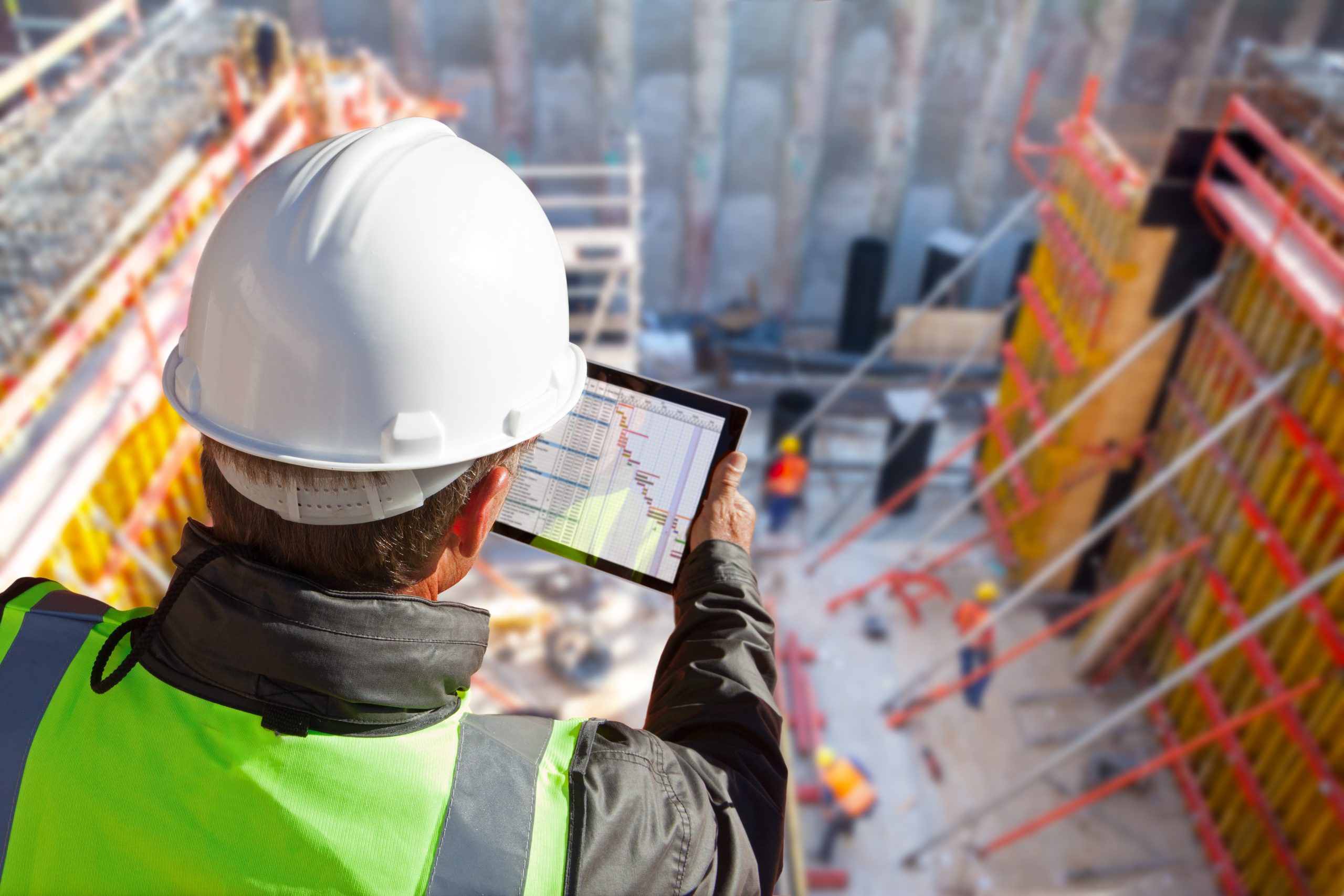 Construction site supervisor reviewing a Gantt chart schedule on a tablet above an active commercial job site