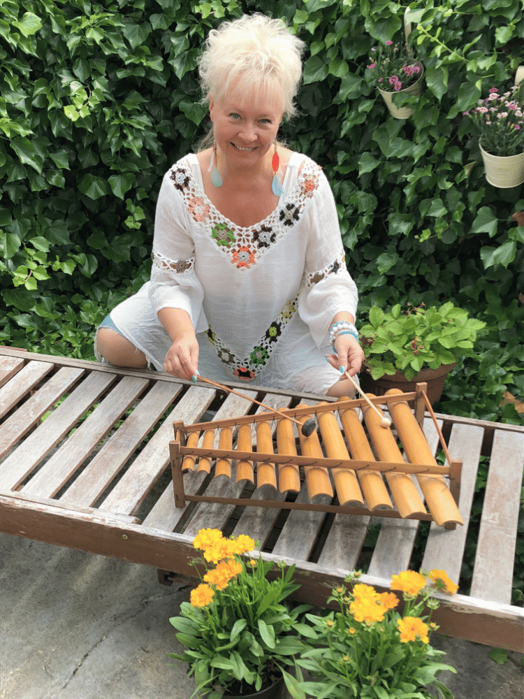 Ania w białej, kwiecistej tunice gra na drewnianym instrumencie perkusyjnym w ogrodzie, otoczona roślinami i zielenią.