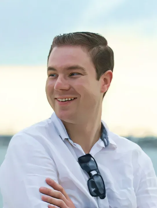 Smiling young man with short brown hair wearing a white shirt and sunglasses hanging from the collar, standing outdoors with arms crossed.